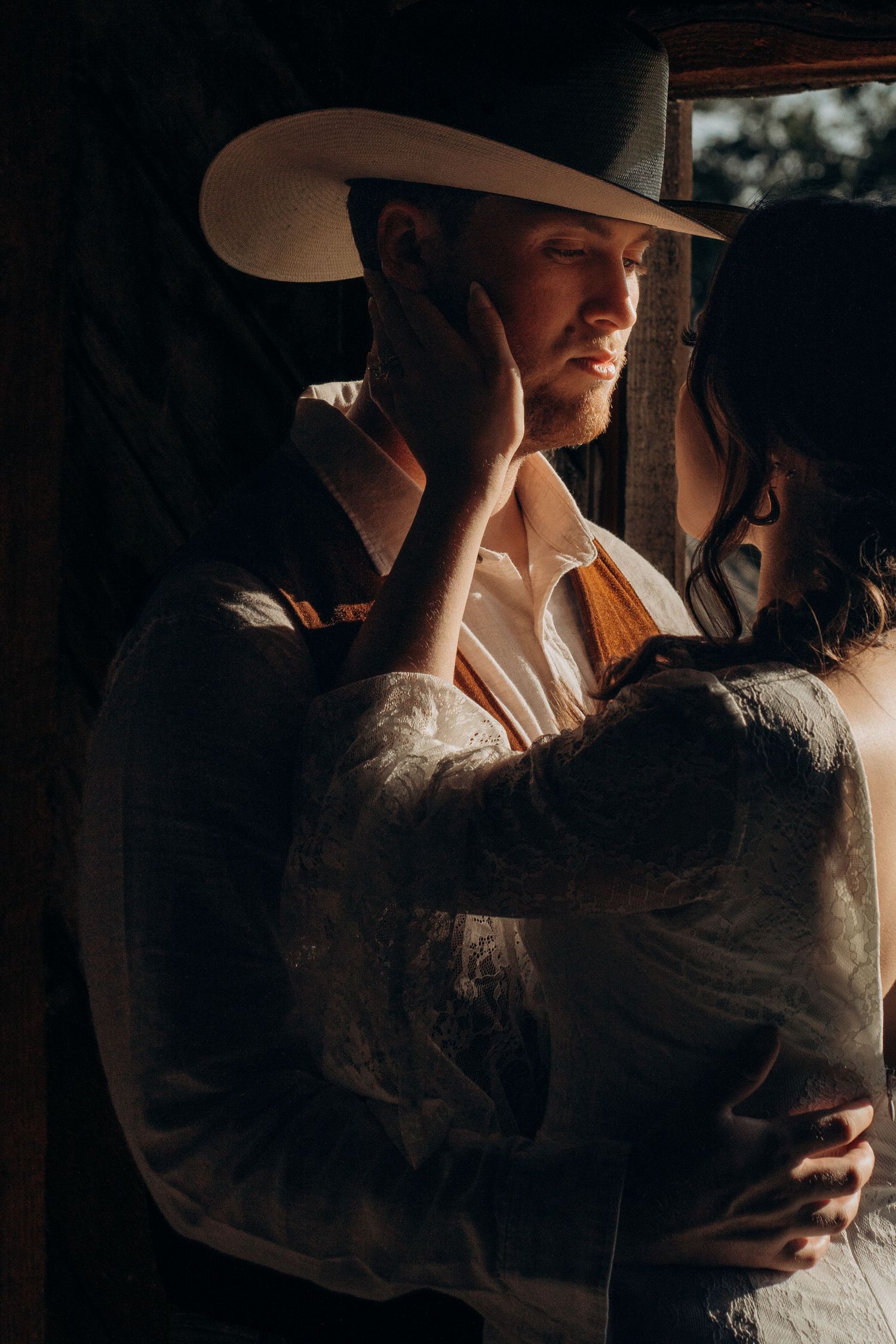 Bride tenderly touches grooms face as he wears a white cowboy hat in warm golden barn light.