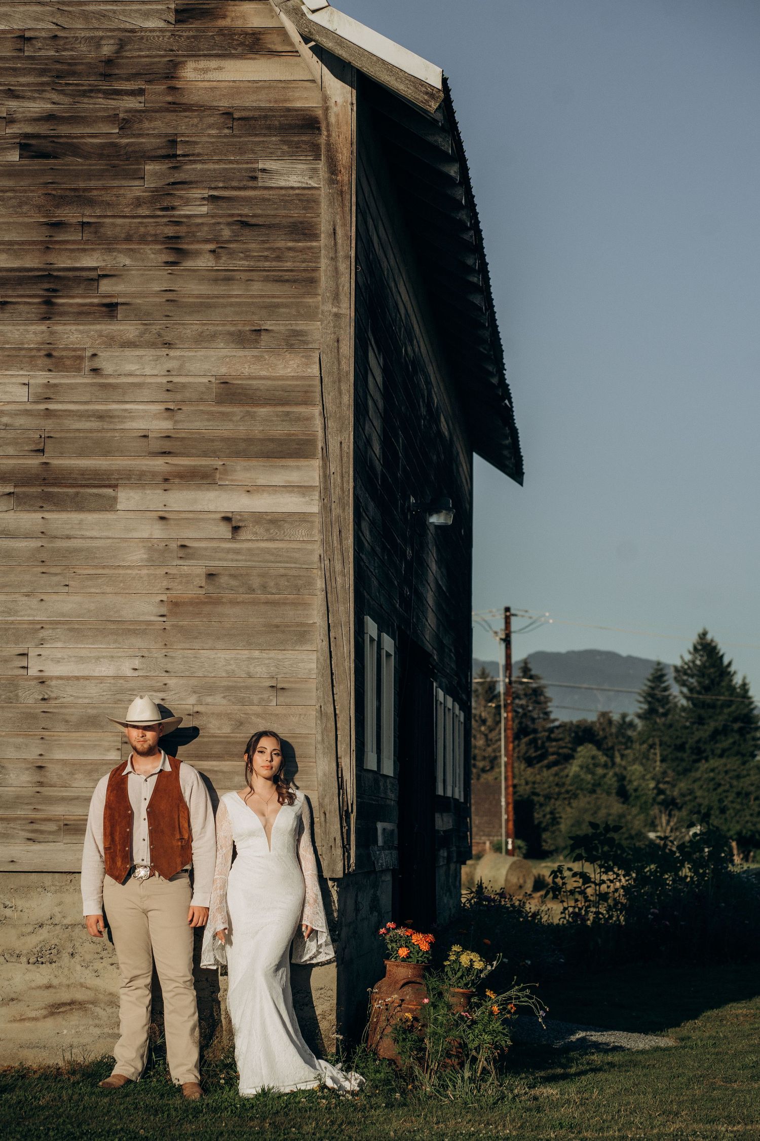 Bride and groom in boho western attire stand beside a large rustic wooden barn under a clear blue sky.