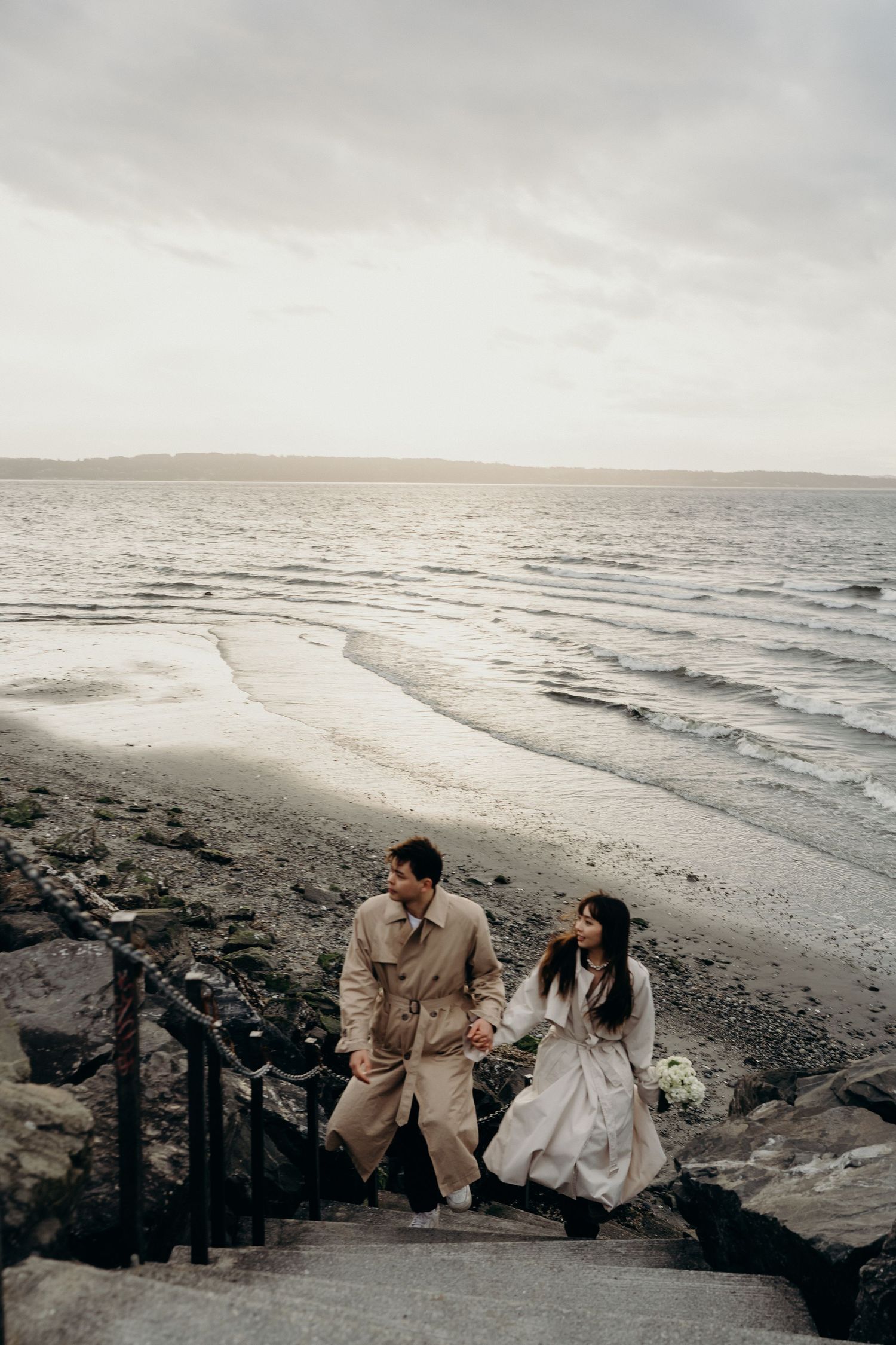 Couple in trench coats holding hands ascending stone steps from overcast ocean beach.