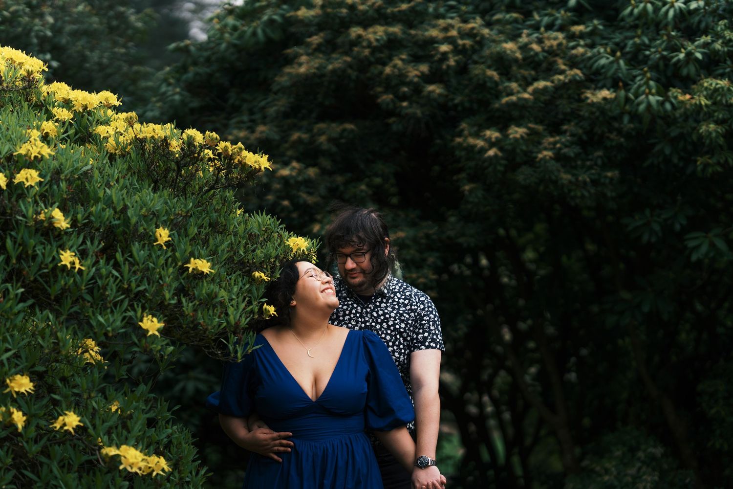 Couple embracing among yellow flowering bushes in a lush green garden during an outdoor engagement session.