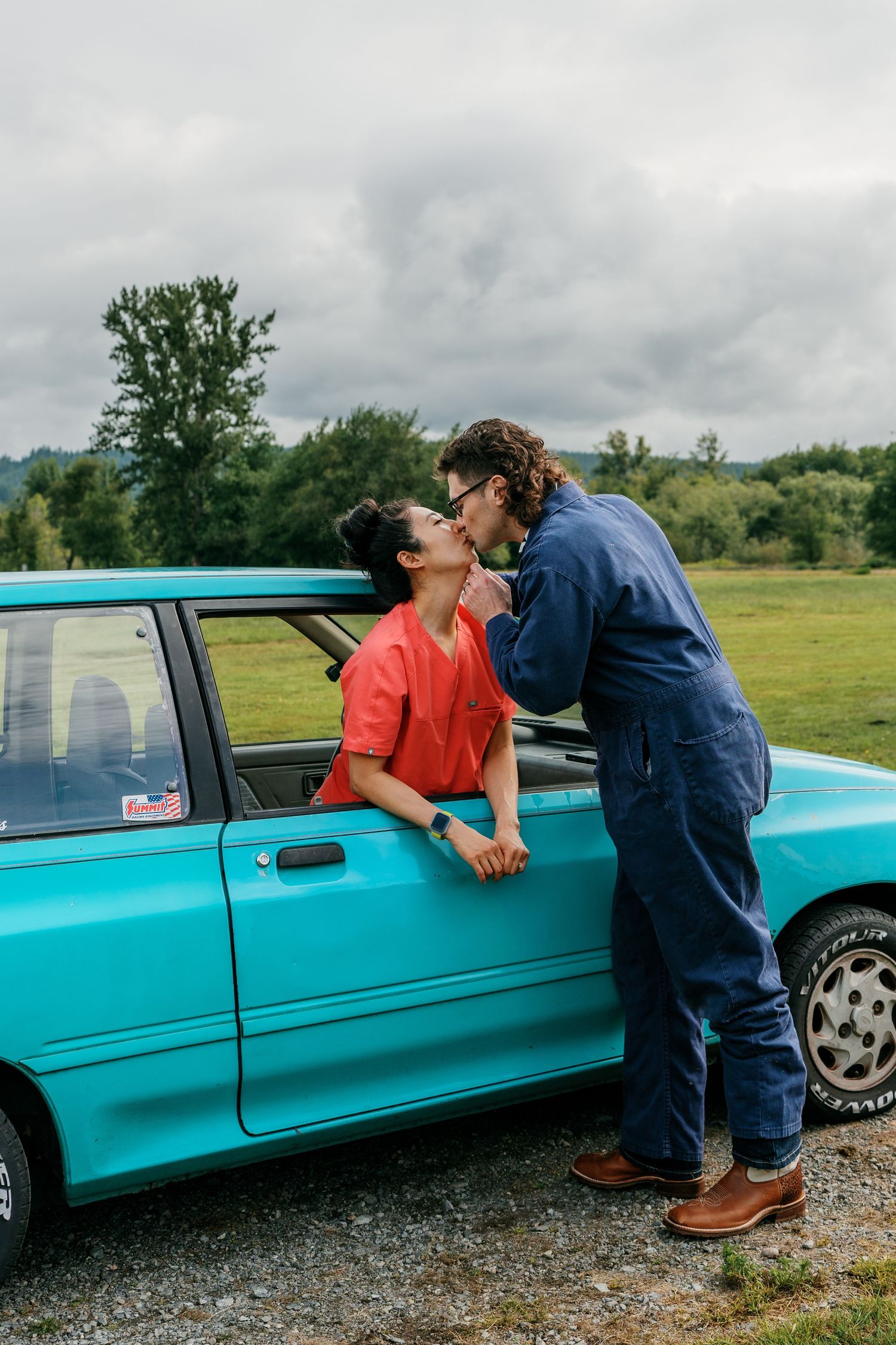 A couple shares a kiss beside a vintage turquoise car in a scenic countryside setting.
