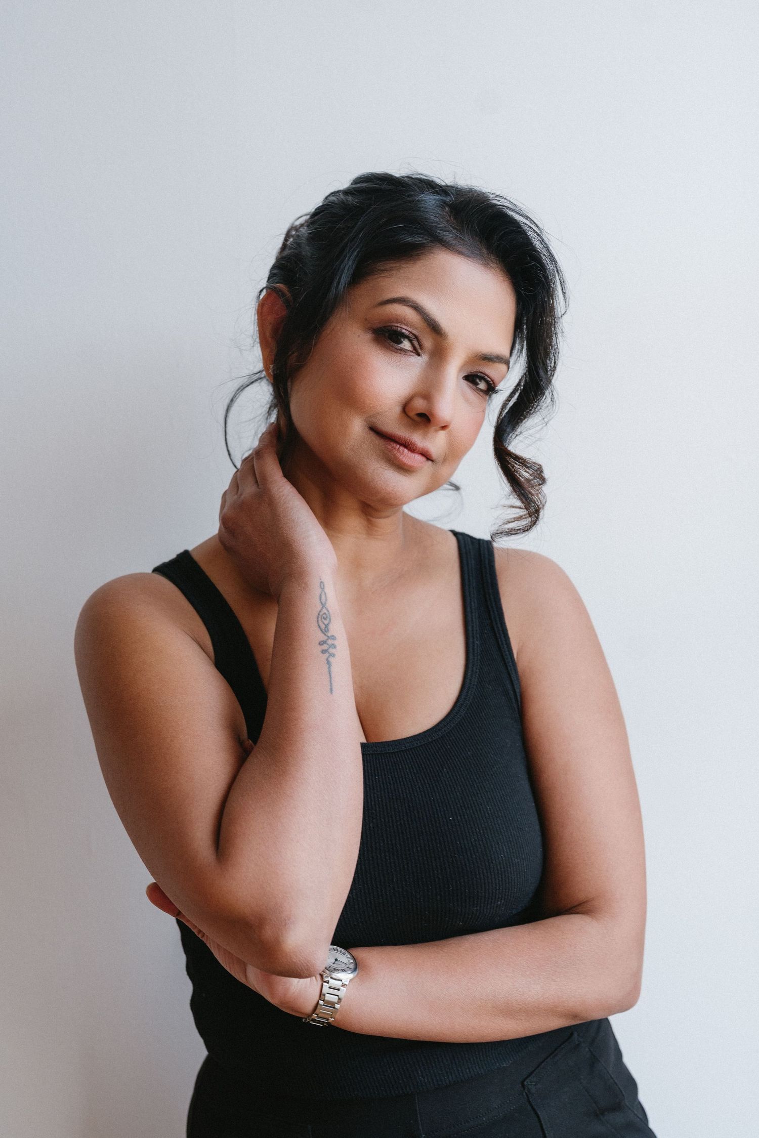 Dark-haired woman in black tank top with wrist tattoo poses gracefully against white wall.