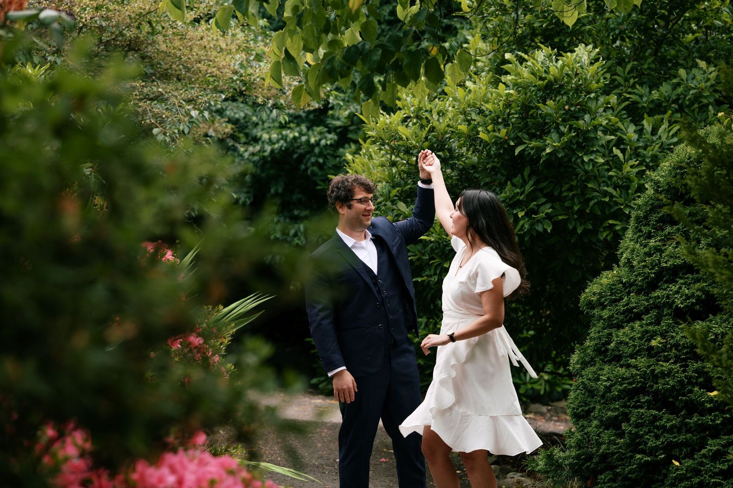 Couple dancing on a garden path, man spinning woman in white dress amid lush green shrubs and pink flowers.
