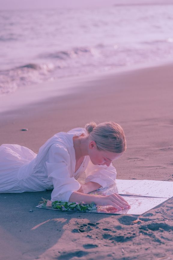 Woman in white dress lying on beach sand writing on paper at sunset.
