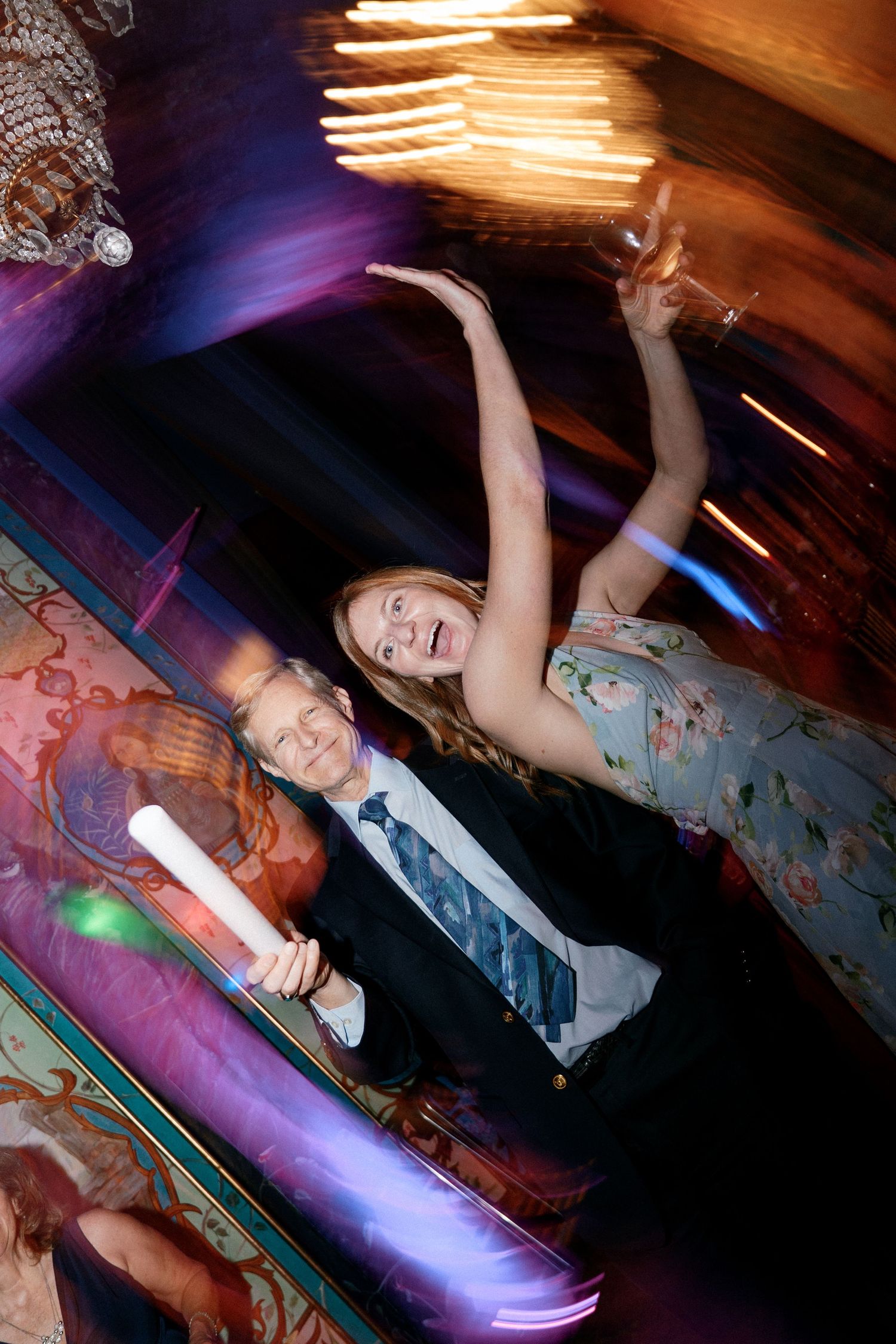 Couple dancing joyfully at a wedding reception under warm chandelier lighting.