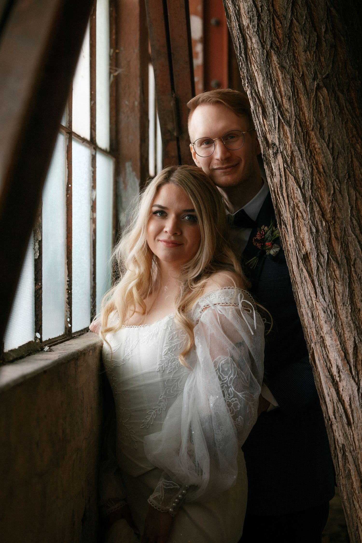 Bride and groom posing by a barred window beside a large tree trunk in a rustic venue.