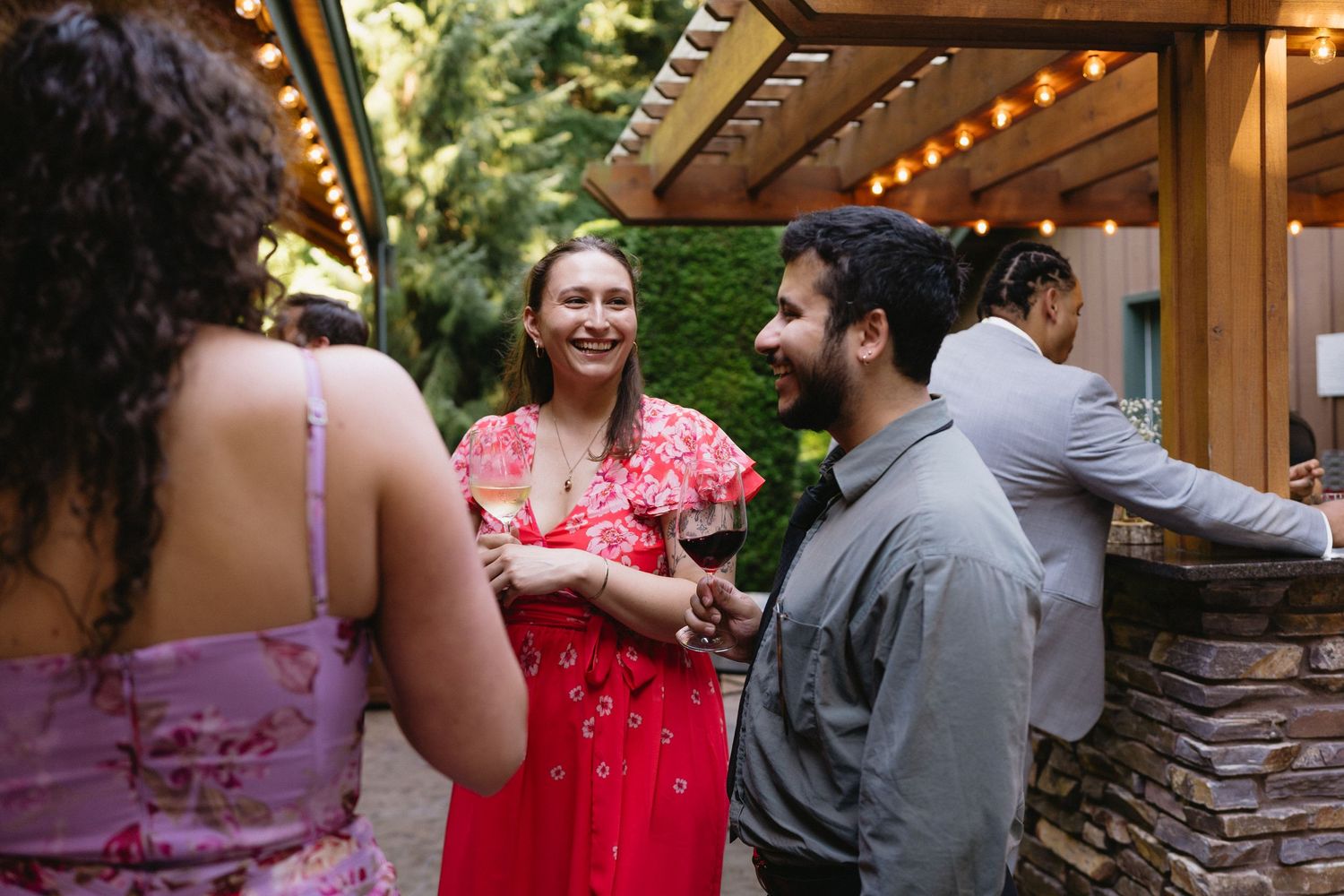Wedding guests laugh and socialize at outdoor reception bar area with string lights.