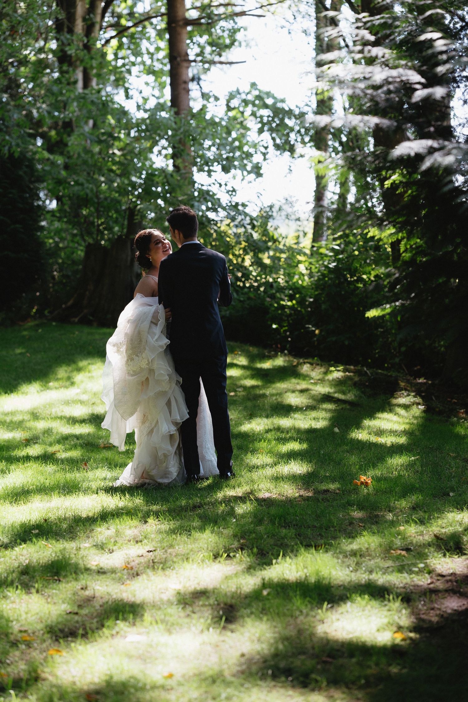 Bride in white gown and groom in navy suit stand together on lush green lawn surrounded by tall trees.