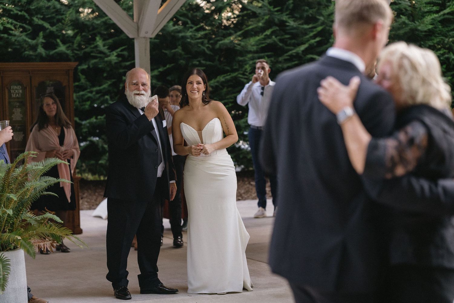 Bride in elegant white gown walks down aisle with father at outdoor wedding ceremony.