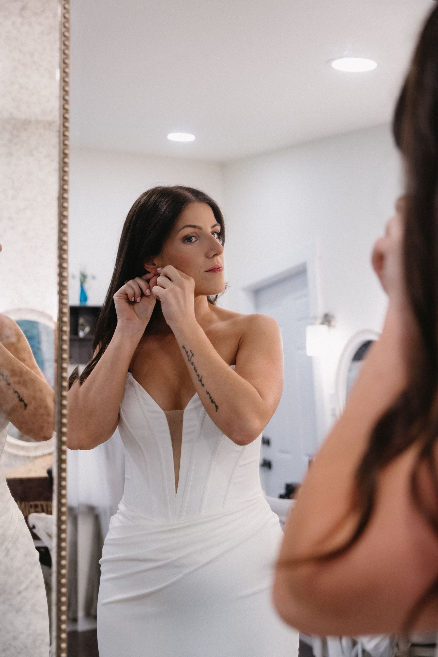 Bride in white strapless corset gown puts on earrings while looking at her reflection in a large mirror.