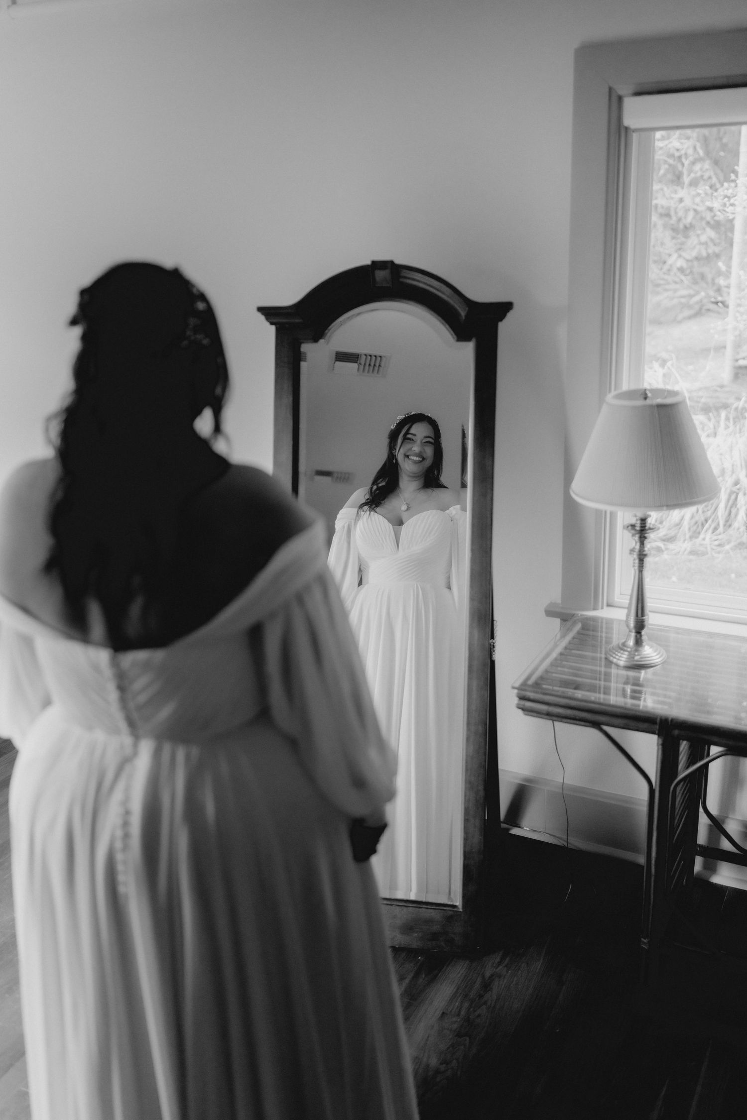 Black and white photo of bride in flowing white gown smiling at her reflection in an ornate full-length mirror.