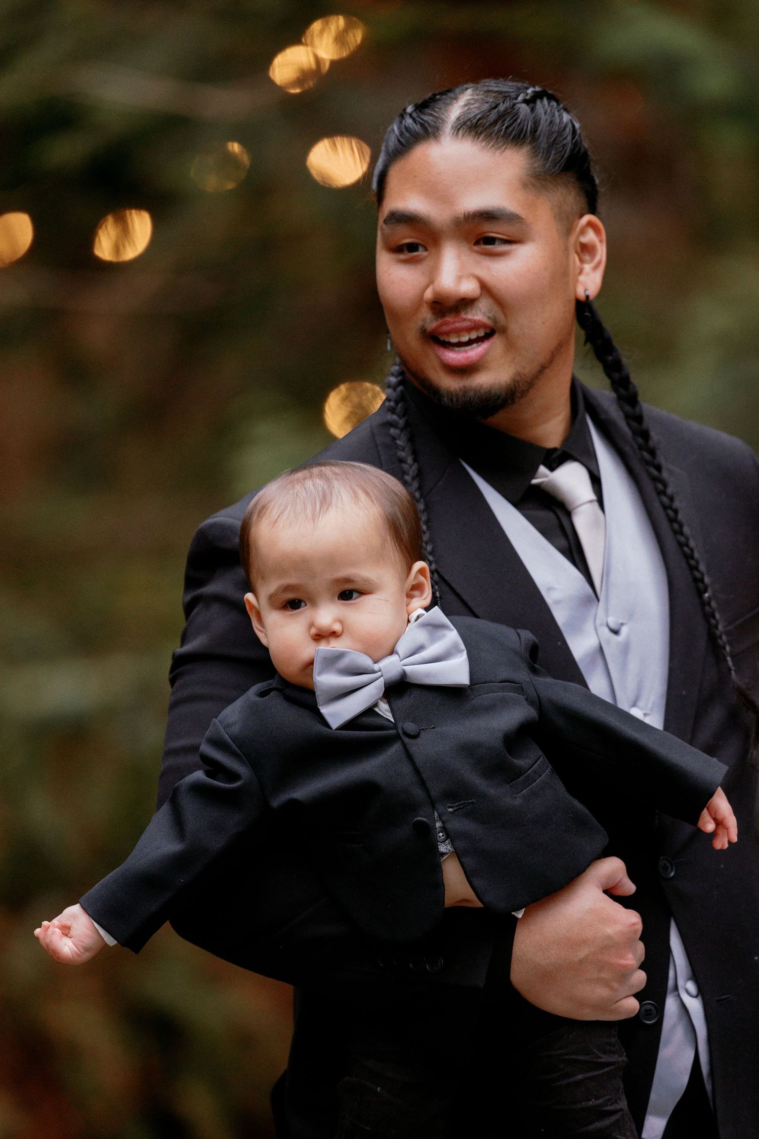 Man with braided hair in black suit holding a baby dressed in matching black tuxedo with bow tie.