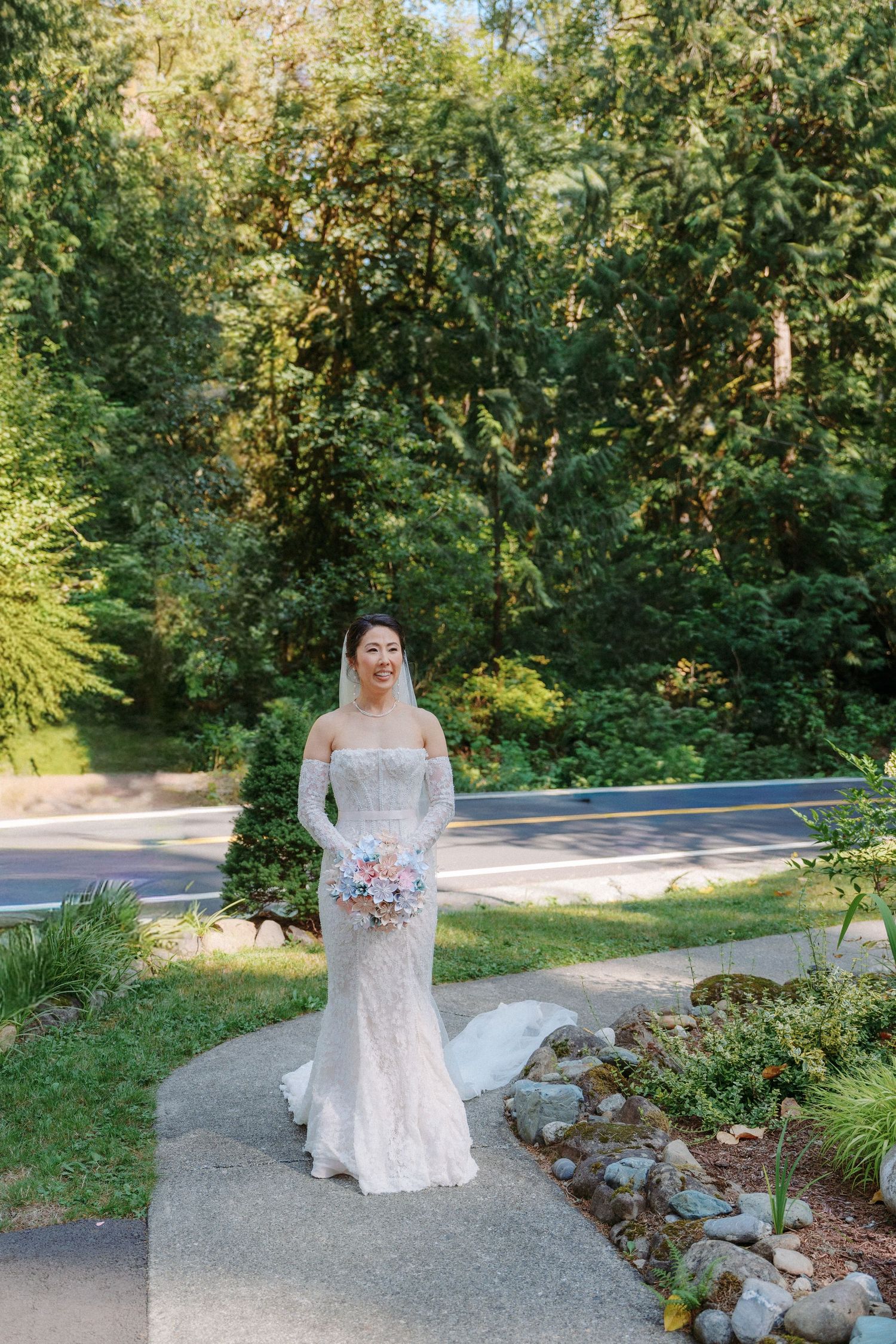 Smiling bride in off-shoulder lace wedding gown holds bouquet on sunlit garden path surrounded by trees.