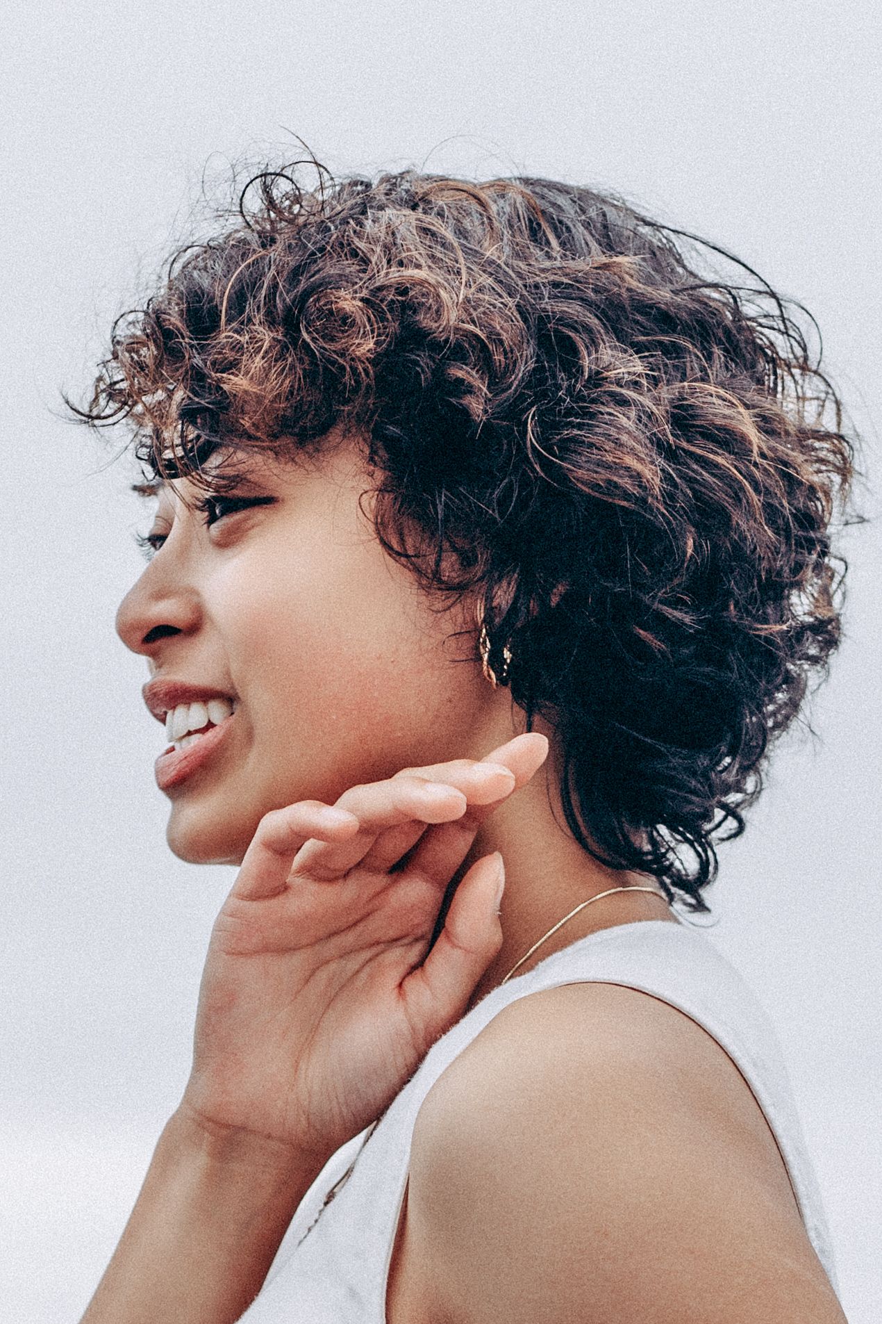 Young woman with short curly hair smiles sideways touching her chin wearing gold necklace.