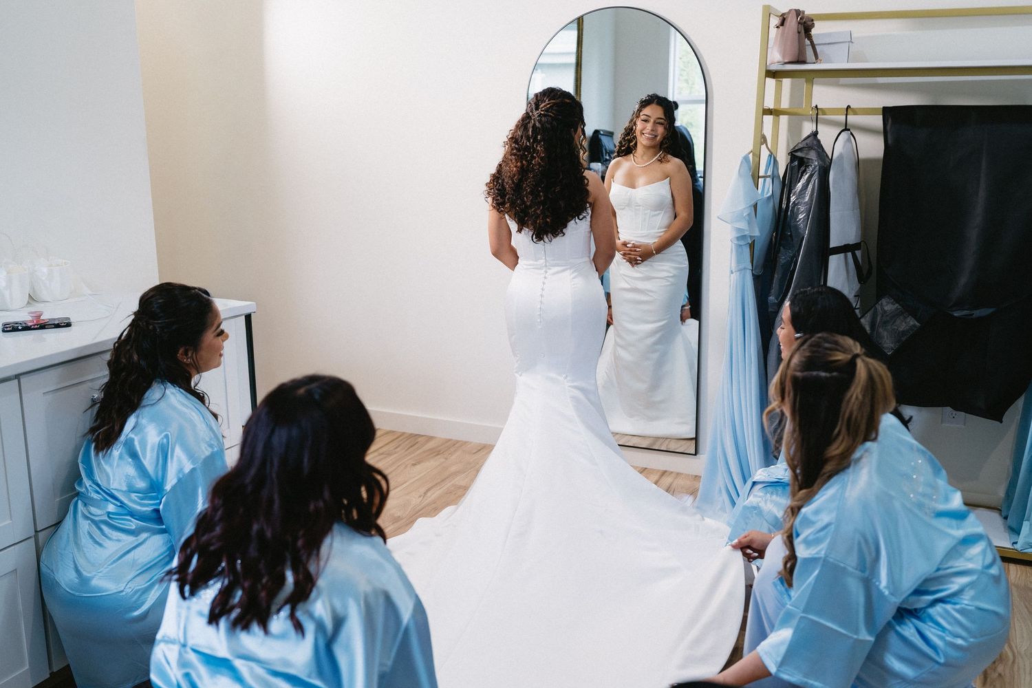 Bridesmaids in blue robes admiring bride in fitted white mermaid gown before full-length mirror.