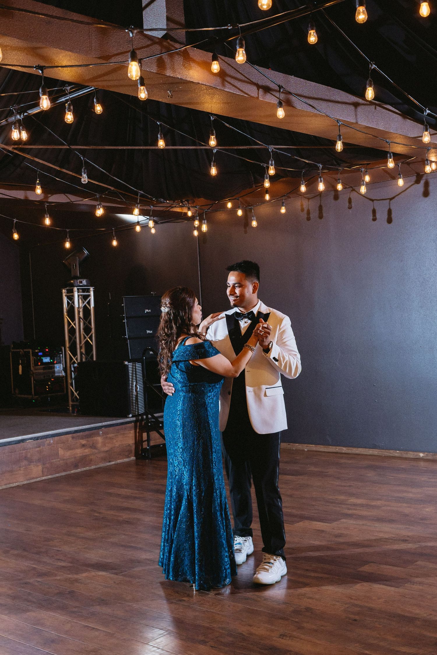 Groom in white jacket sharing mother-son dance under vintage string lights.