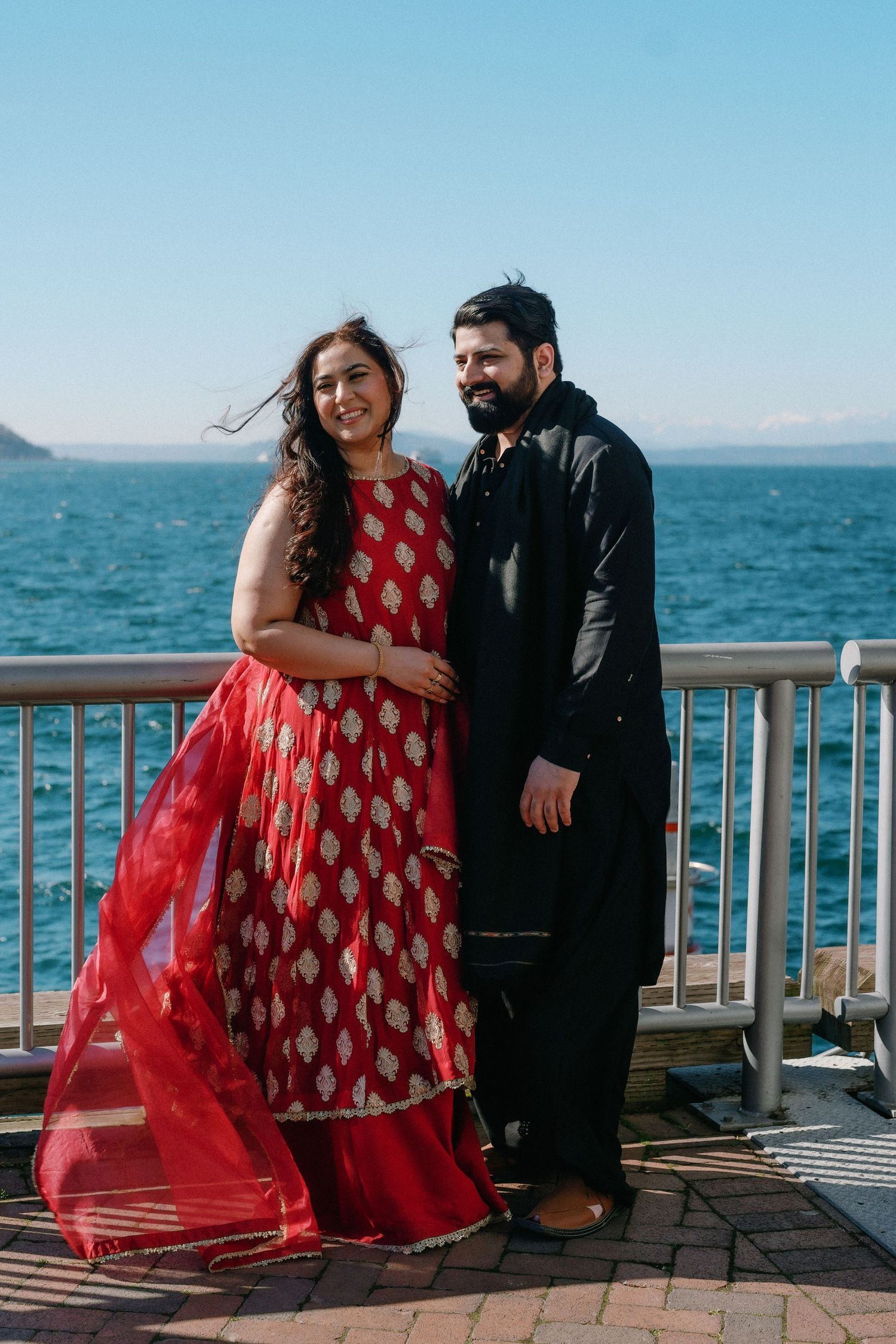 Couple posing by waterfront railing, woman in red Indian dress and man in black kurta, smiling at camera.