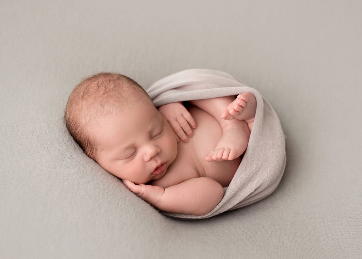 Newborn baby swaddled on back with tiny hands and feet showing against grey backdrop, Victoria BC photography