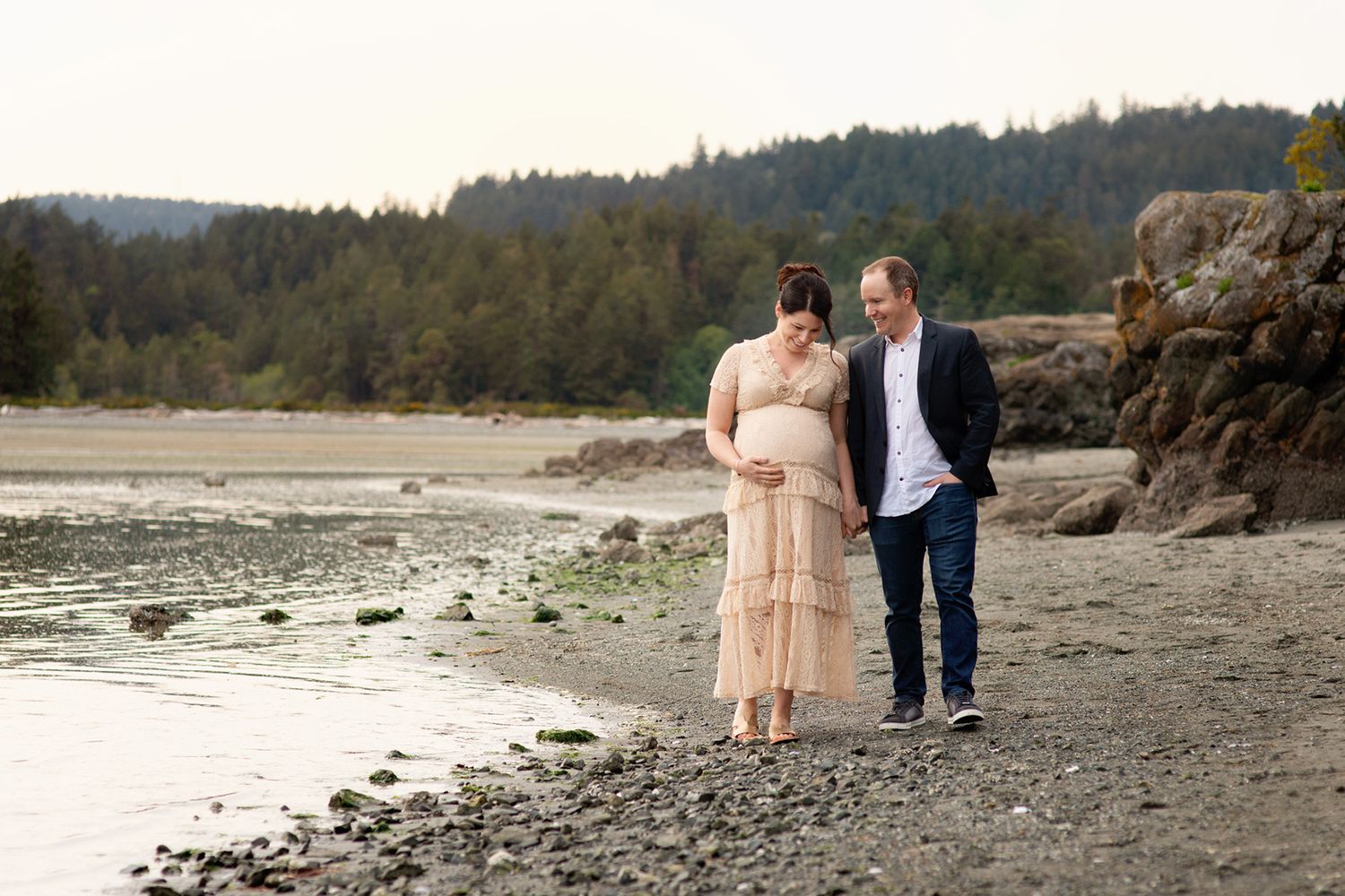 An expecting couple walks together on a rocky beach at sunset with forest and cliffs in the background, maternity photography, Victoria BC