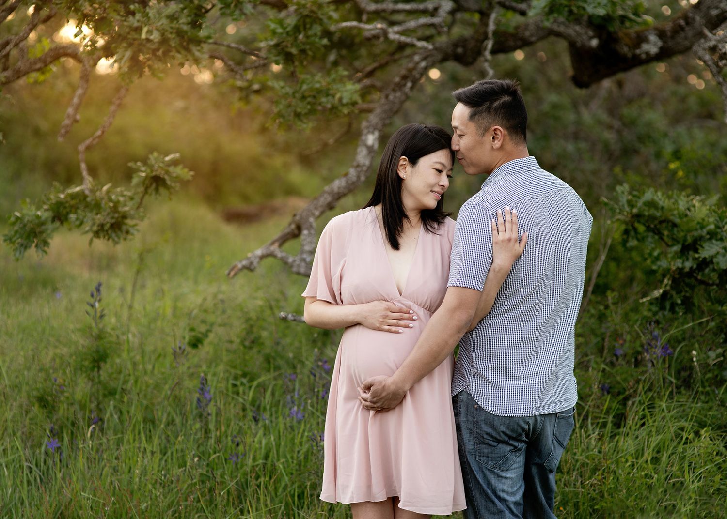 An excepting couple shares a tender moment together in a sunlit meadow with wildflowers and trees in the background.