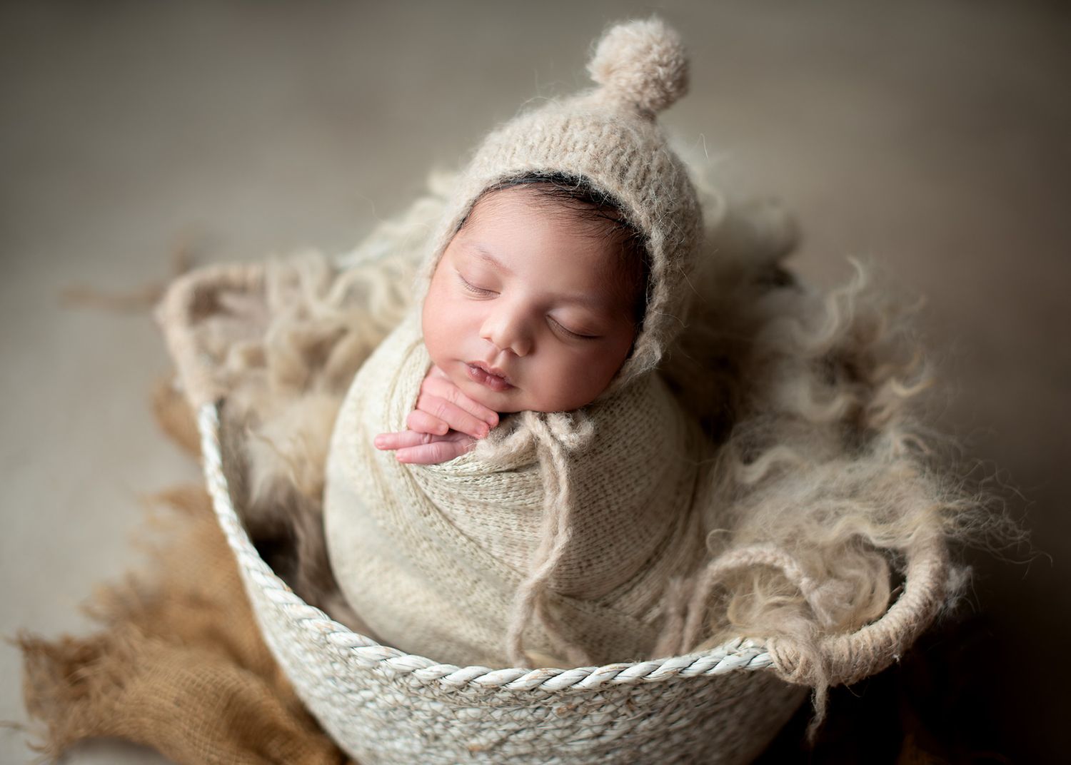 Sleeping newborn wrapped in beige swaddle wearing white knit hat rests peacefully in textured bowl on fuzzy blanket, Victoria BC