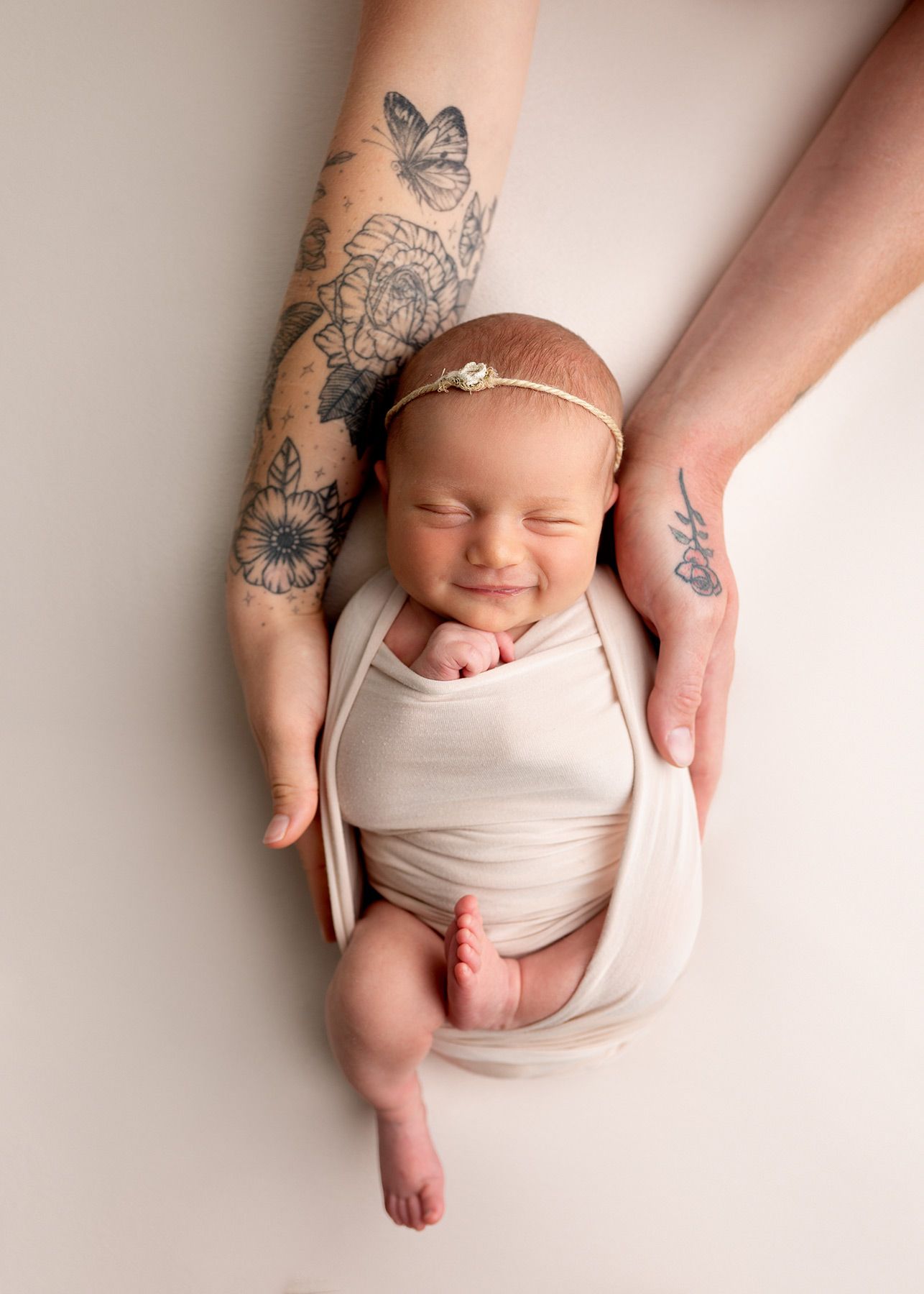 A happy sleeping newborn baby wrapped in beige fabric is cradled in tattooed hands of parents, Victoria BC newborn photography