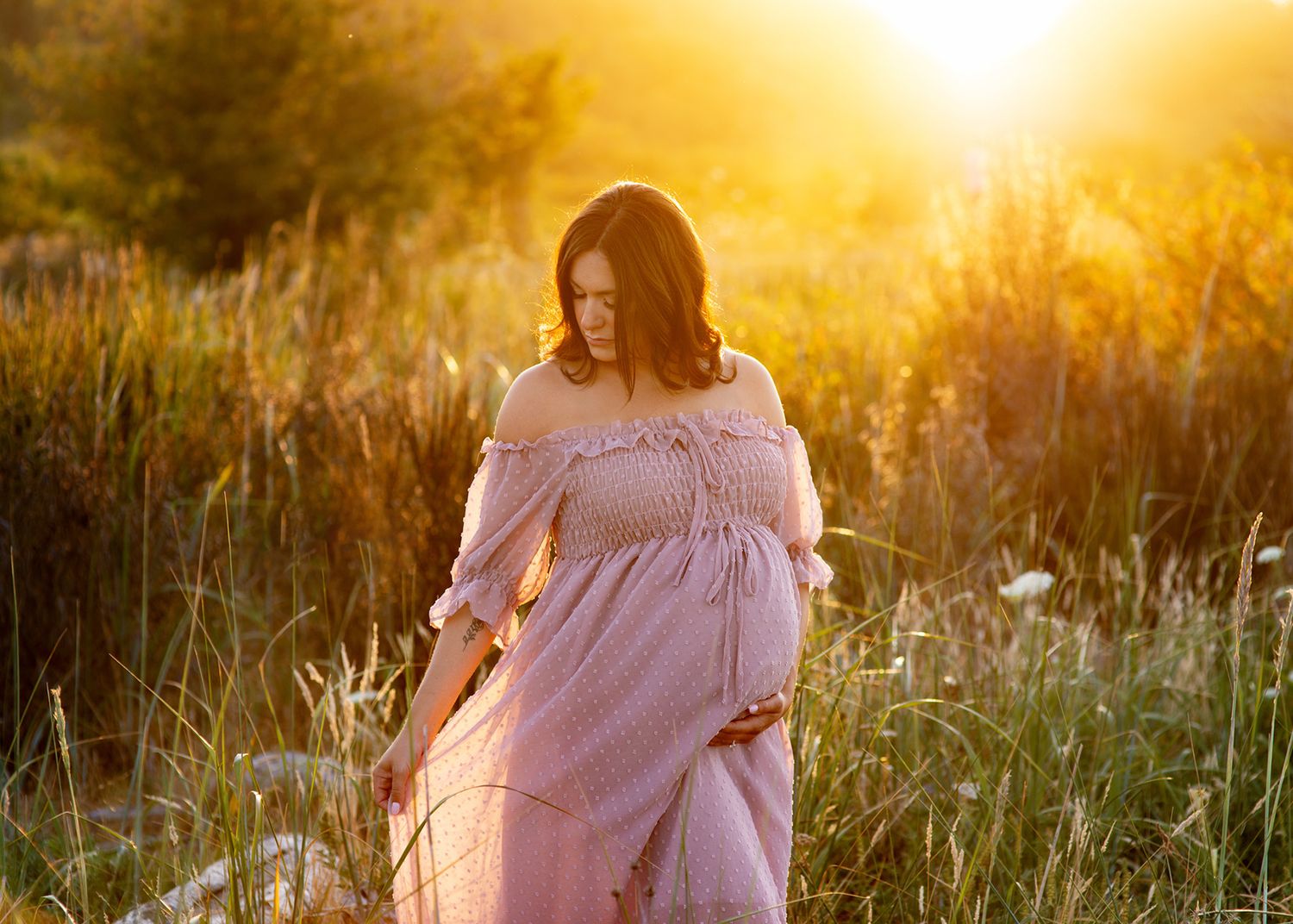 An excepting mother in a flowing pink dress stands in a golden sunlit field of tall grass during sunset, Victoria BC photography