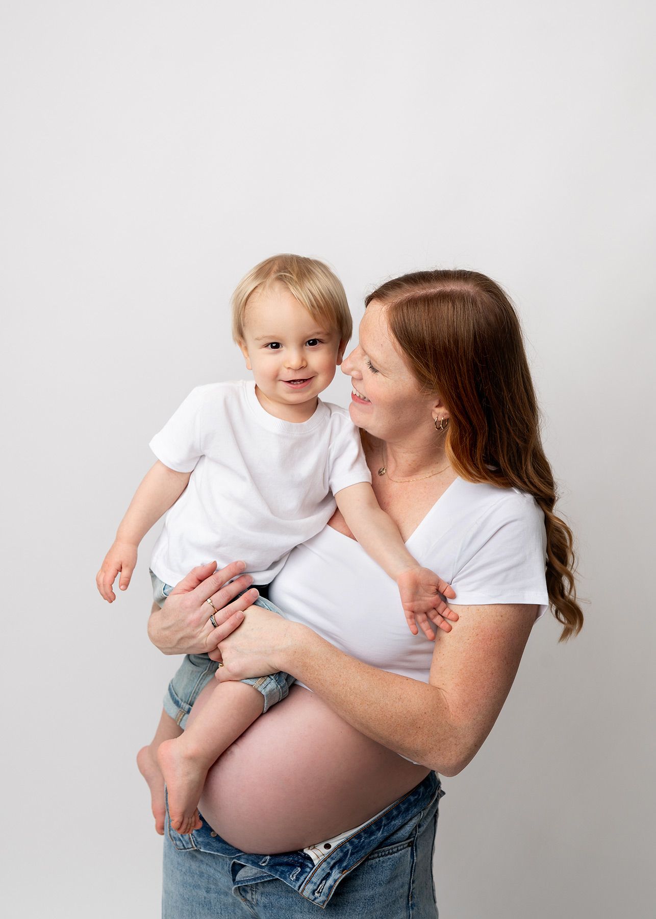 A pregnant woman in white shirt and jeans holds and interacts with a toddler against a light background, Victoria BC studio maternity photo