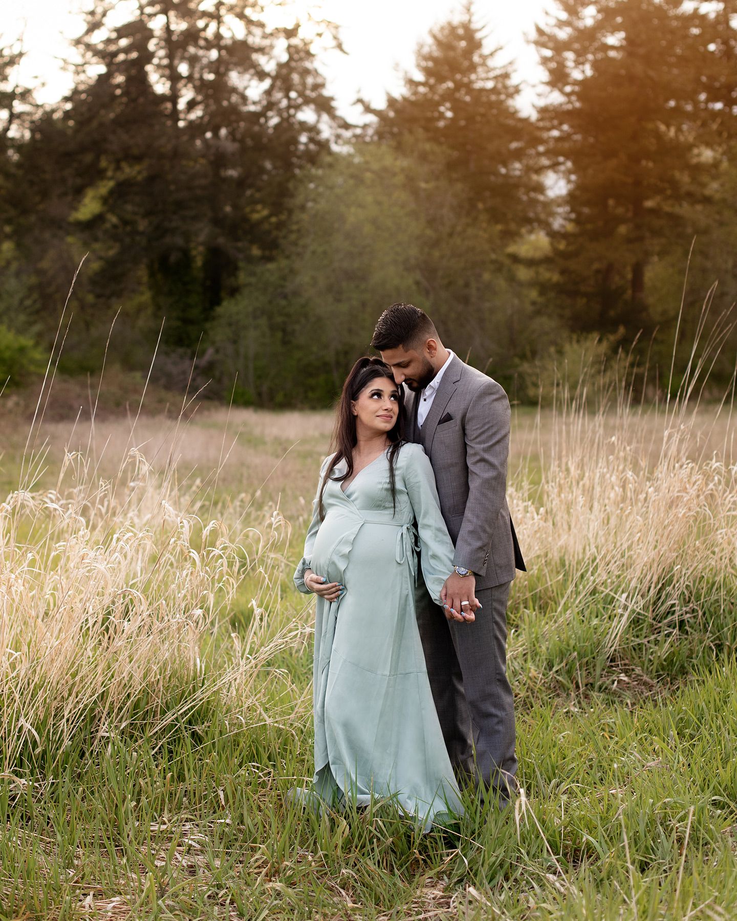 An expecting couple in formal attire embraces tenderly in a serene field of tall grass during golden hour, Victoria BC