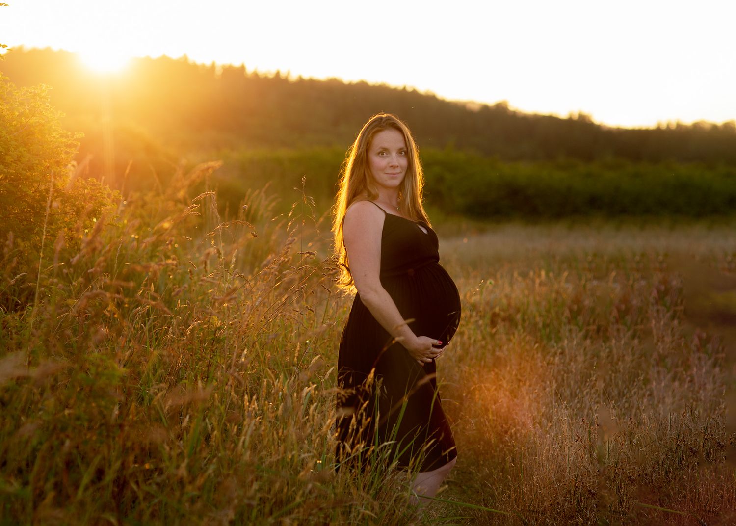 A golden sunset illuminates a grassy meadow as pregnant mother stands silhouetted against the warm evening light, Victoria BC