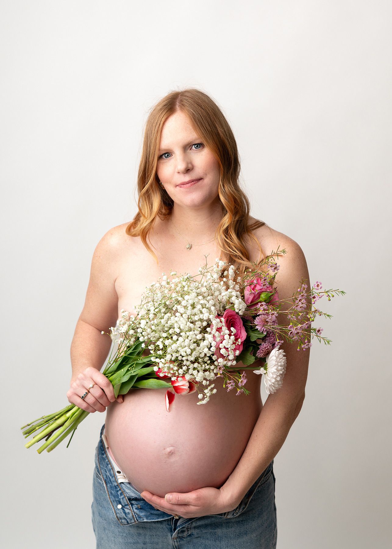 Maternity photo series showing pregnant woman in jeans and white top holding baby's breath flowers against white backdrop.