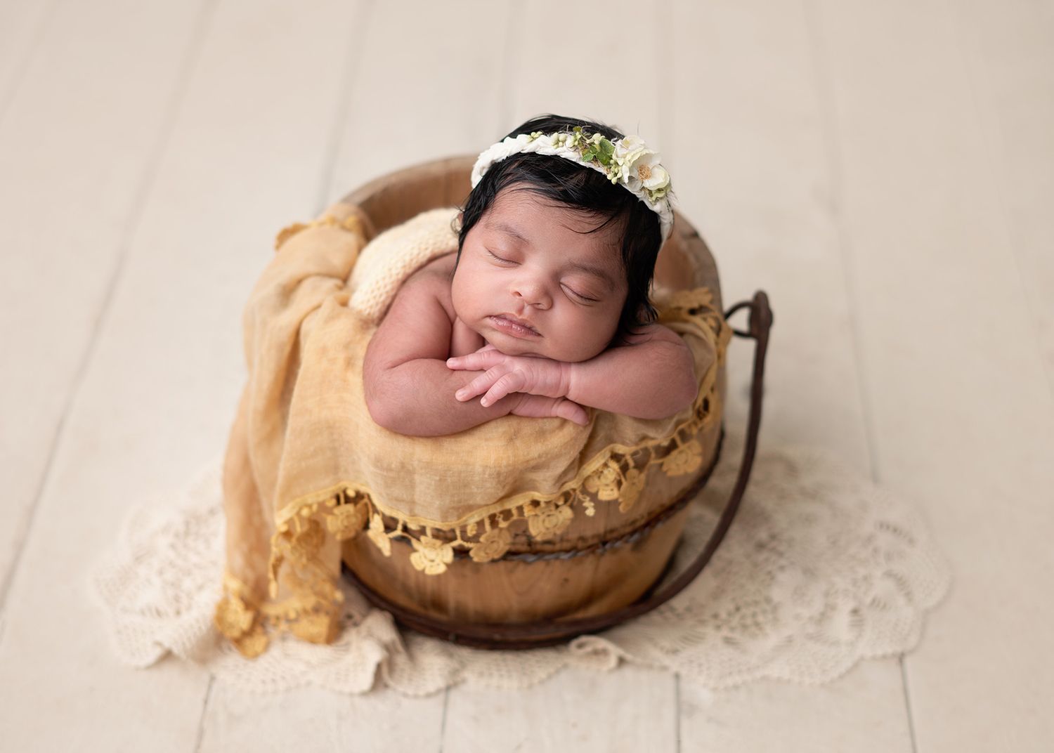 Sleeping newborn in a floral headband rests peacefully in a wooden bowl with soft yellow fabric and lace backdrop, Victoria BC newborn photo