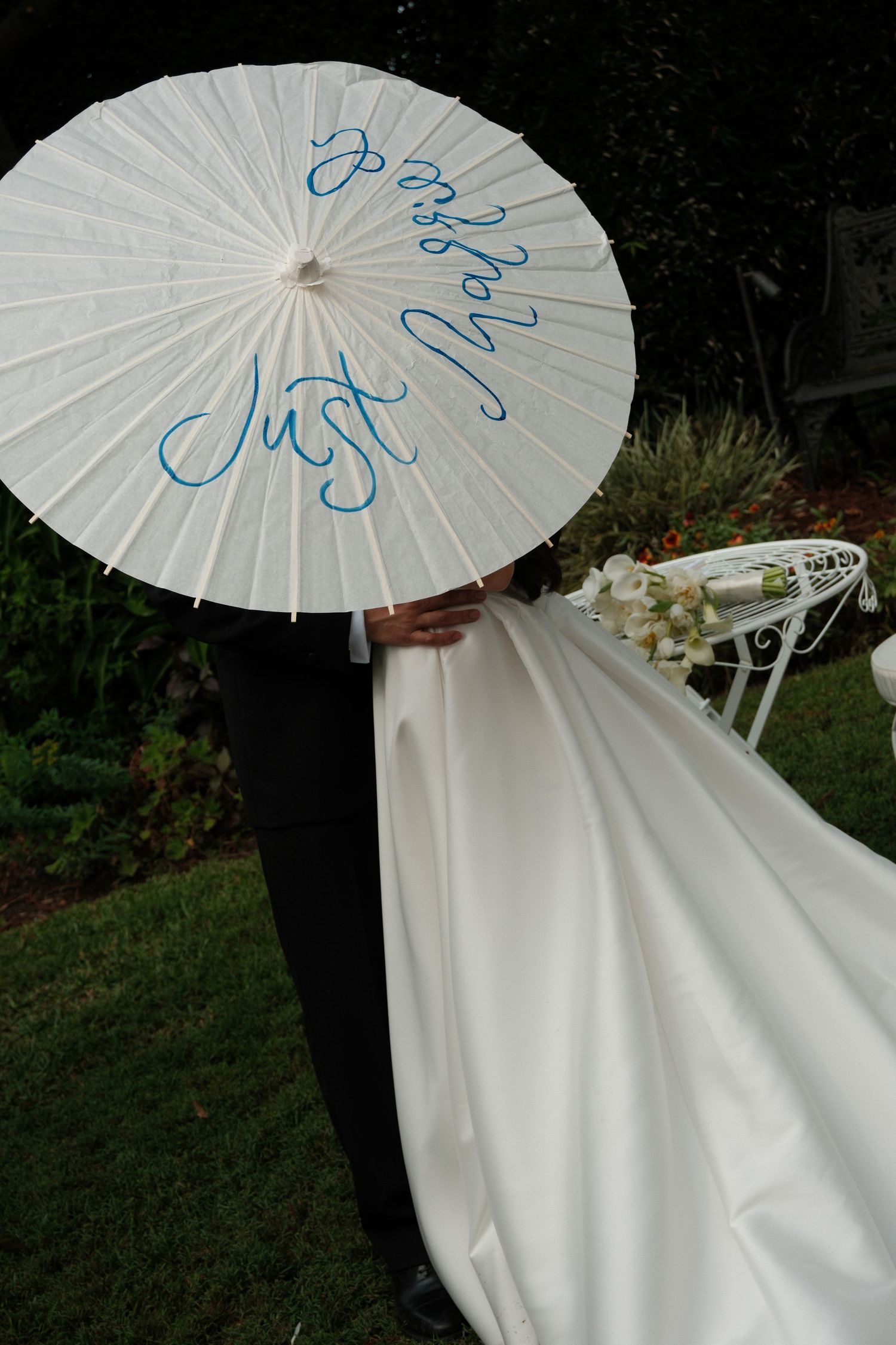 A white parasol with blue calligraphy writing is held over a flowing white wedding dress in an outdoor garden setting.