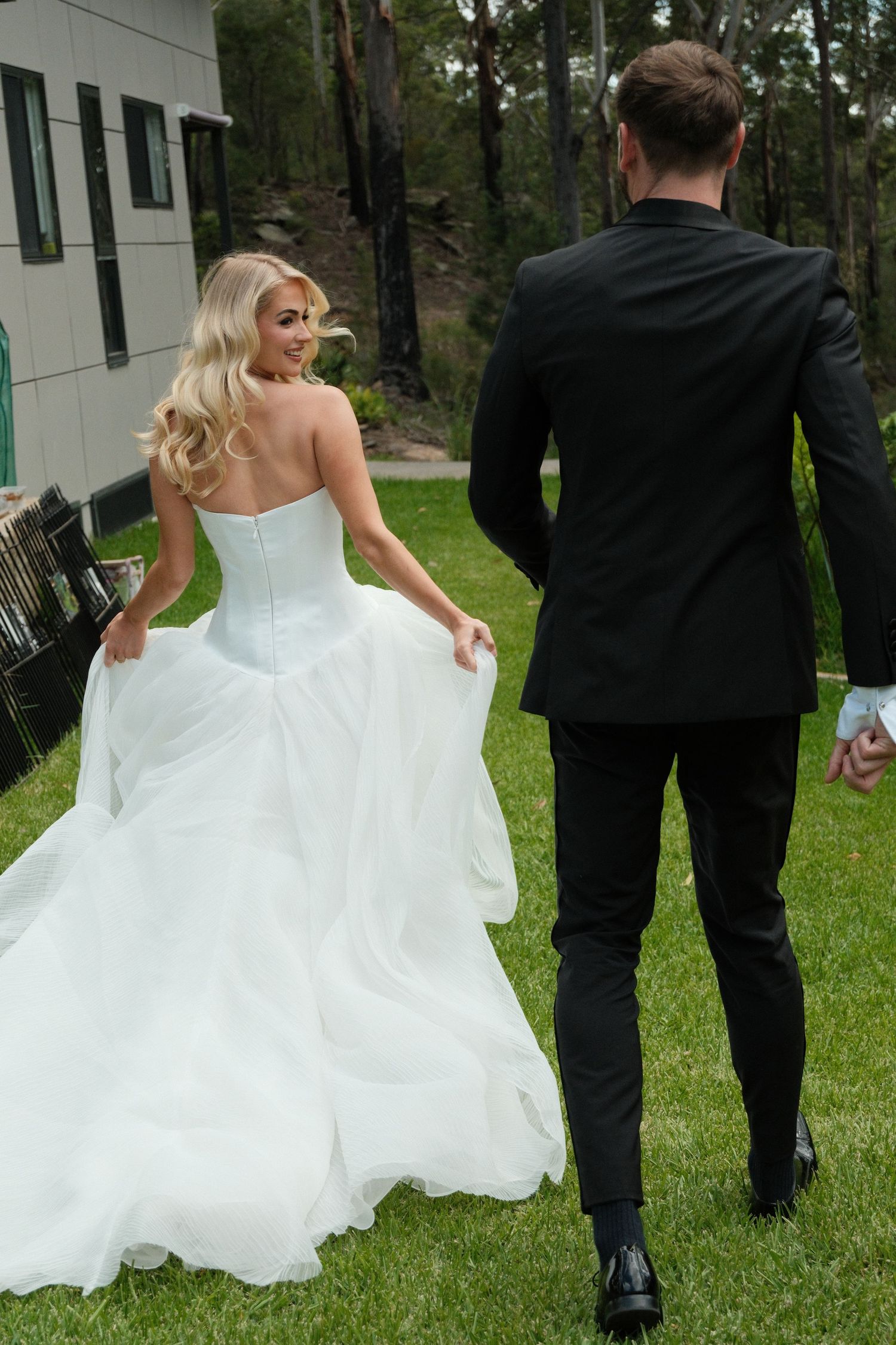 A couple in wedding attire walks through a grassy outdoor setting with tall trees in the background.
