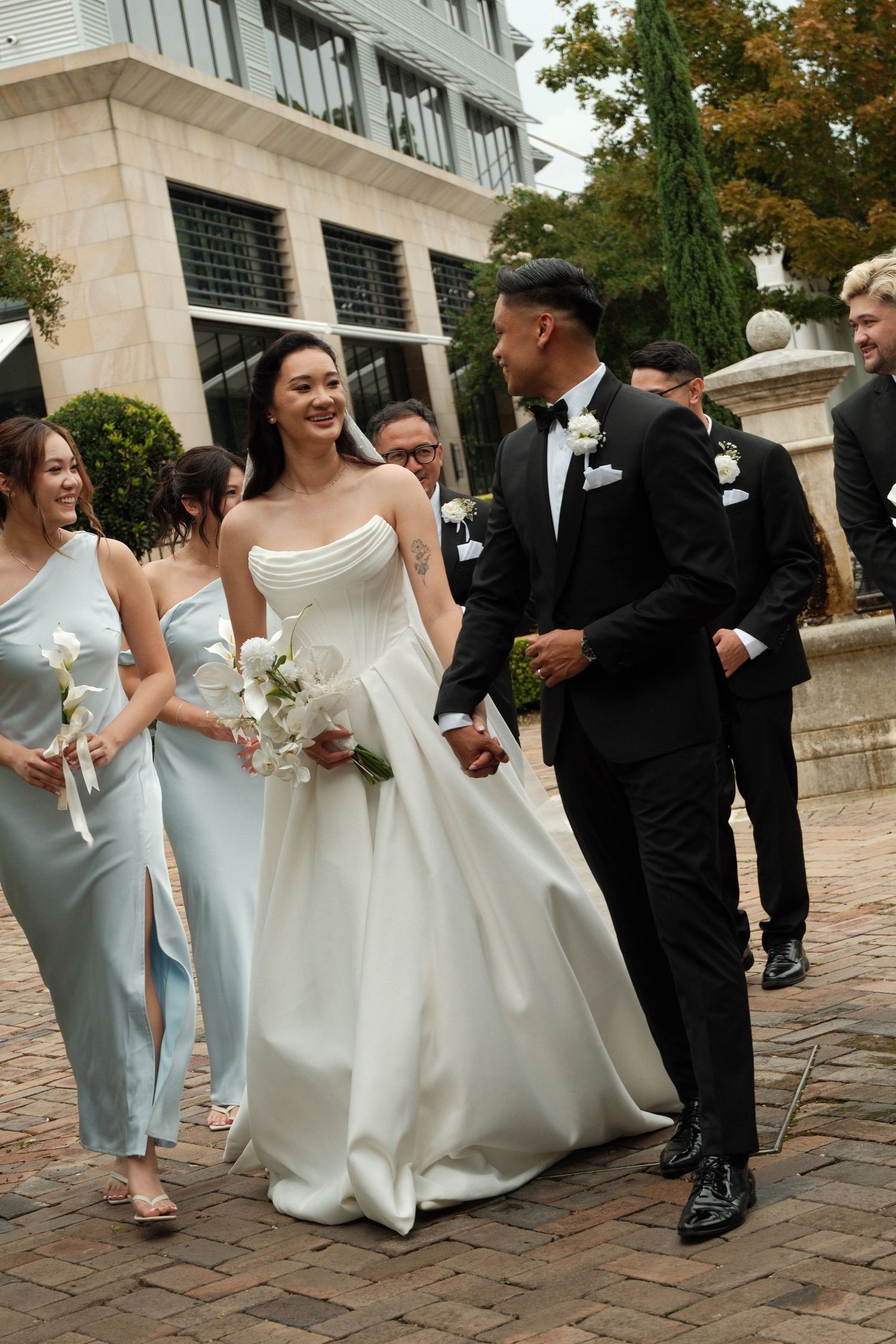 Wedding party walks arm in arm along a brick pathway outside a stone building in light blue and black attire.