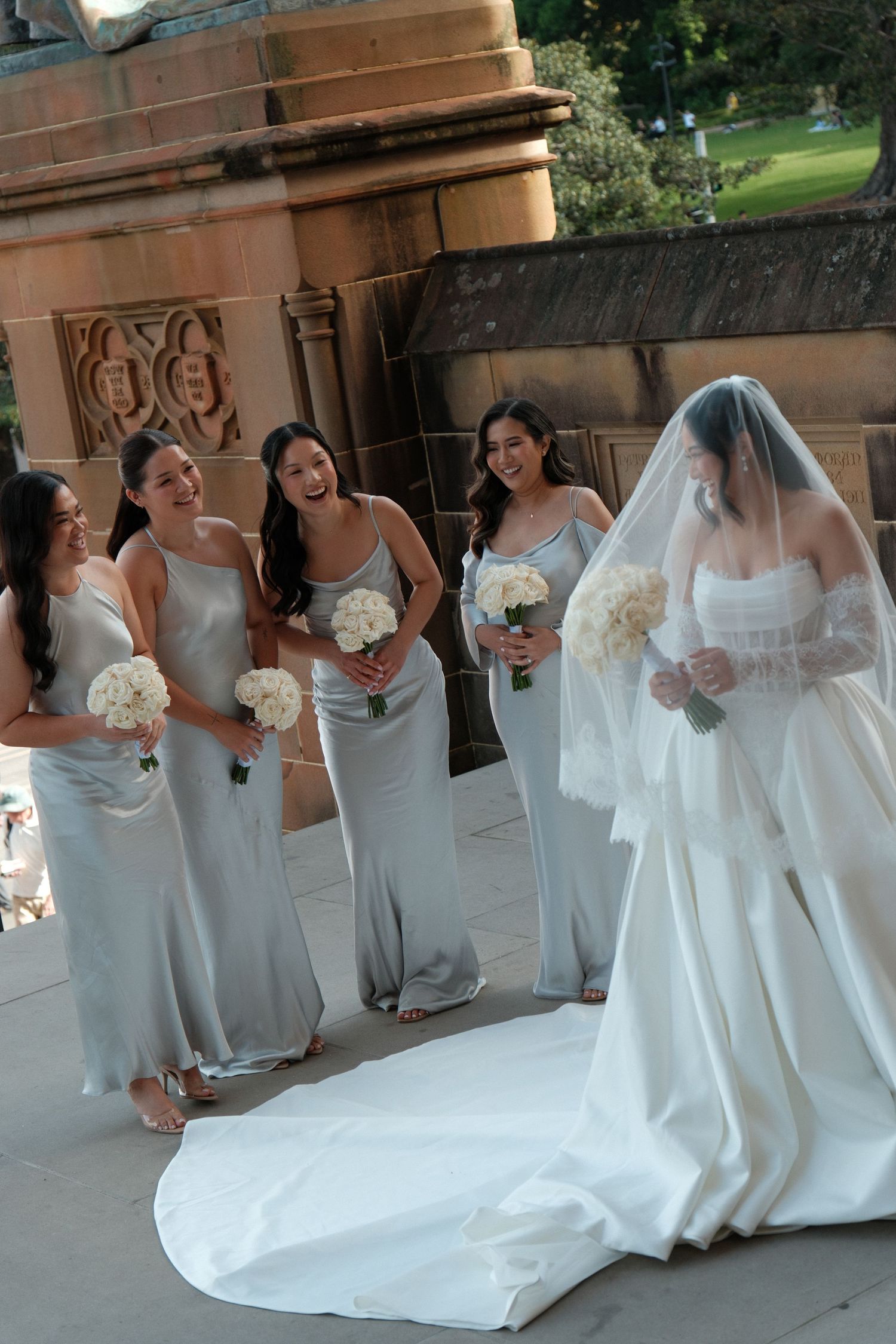 Bride in white gown and veil laughing with four bridesmaids in silver dresses holding white bouquets outside stone venue.