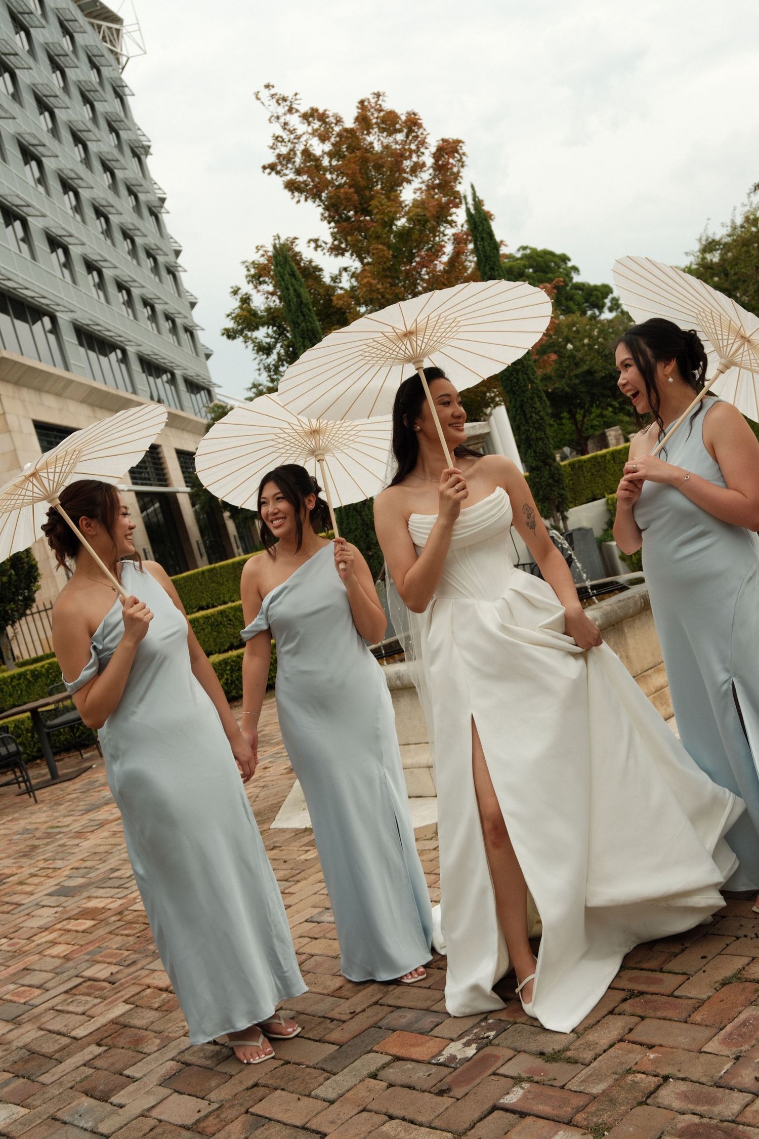 Bridesmaids in pale blue dresses holding white paper parasols pose with bride in elegant white gown on brick patio.