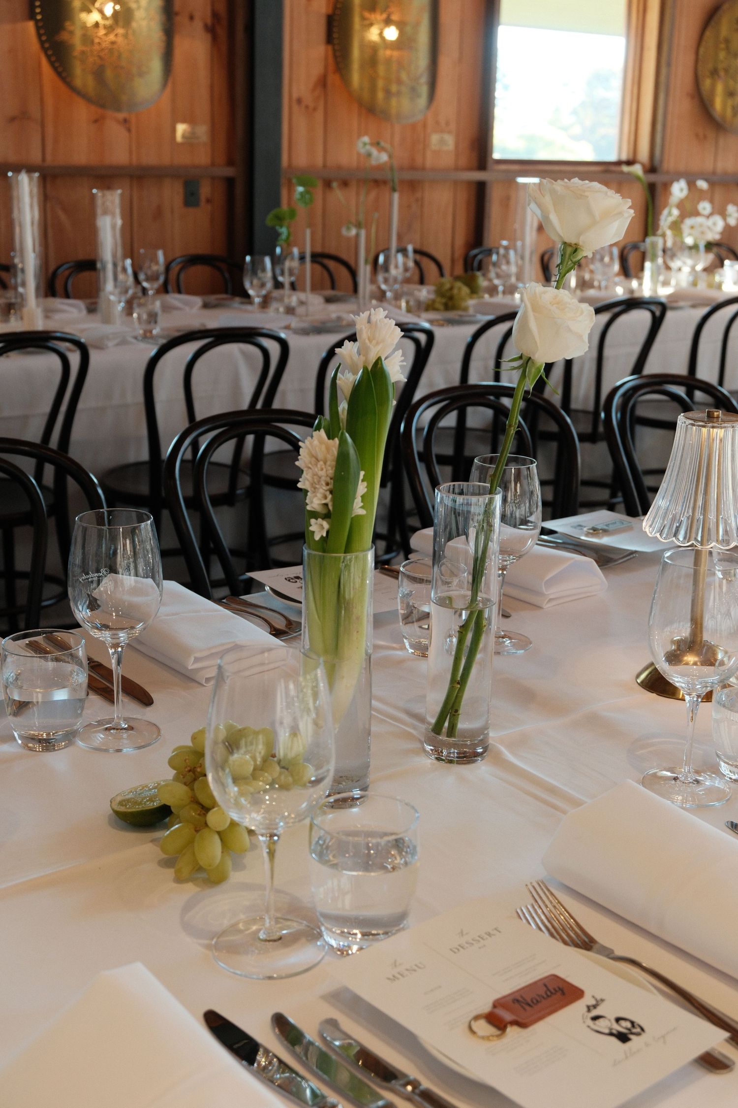 Elegant wedding reception table setting with white flowers in glass vases, wine glasses, and white linens in a warm wooden venue.