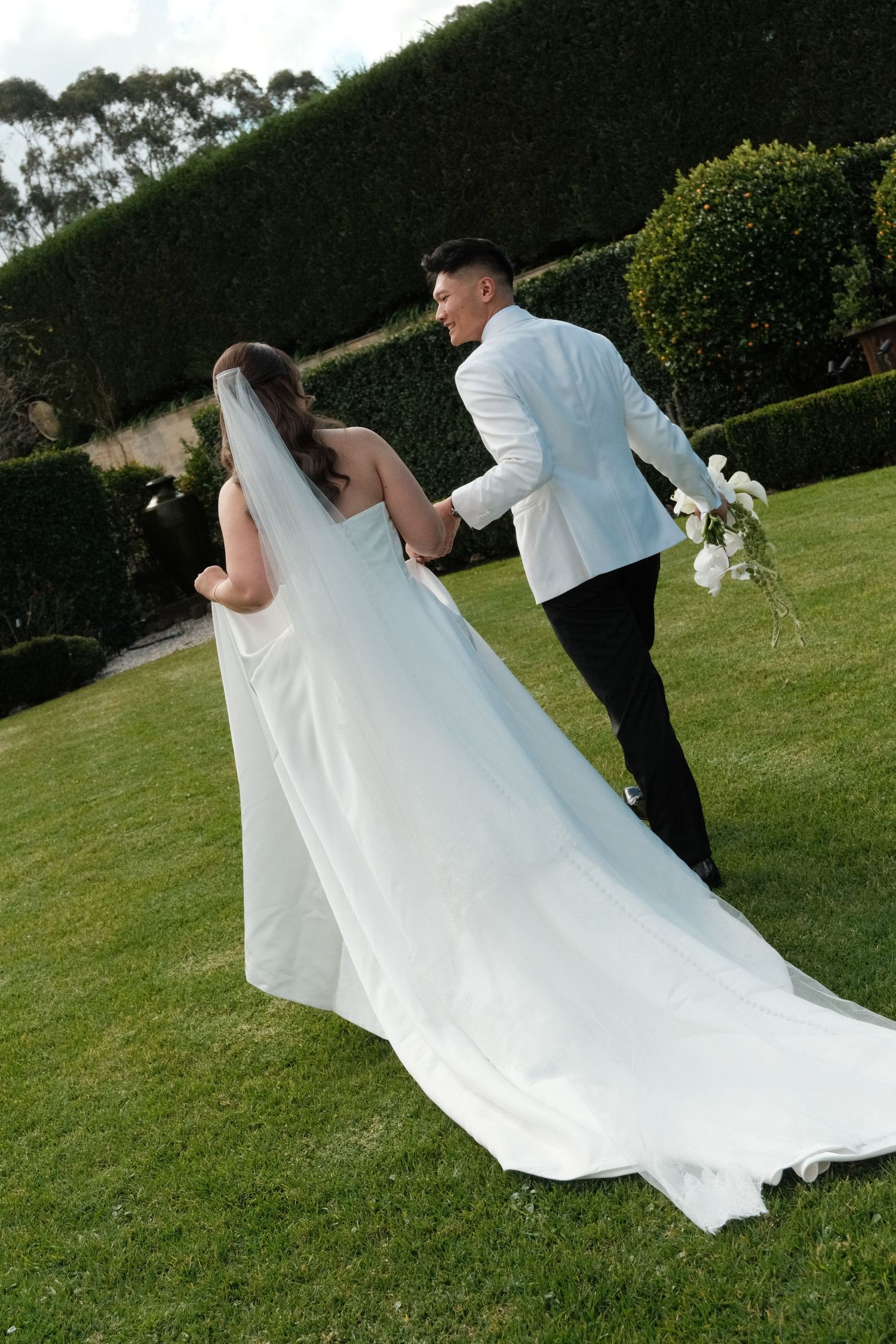 Bride in white gown with veil and groom in white suit holding flowers walk hand in hand on lush green garden lawn.