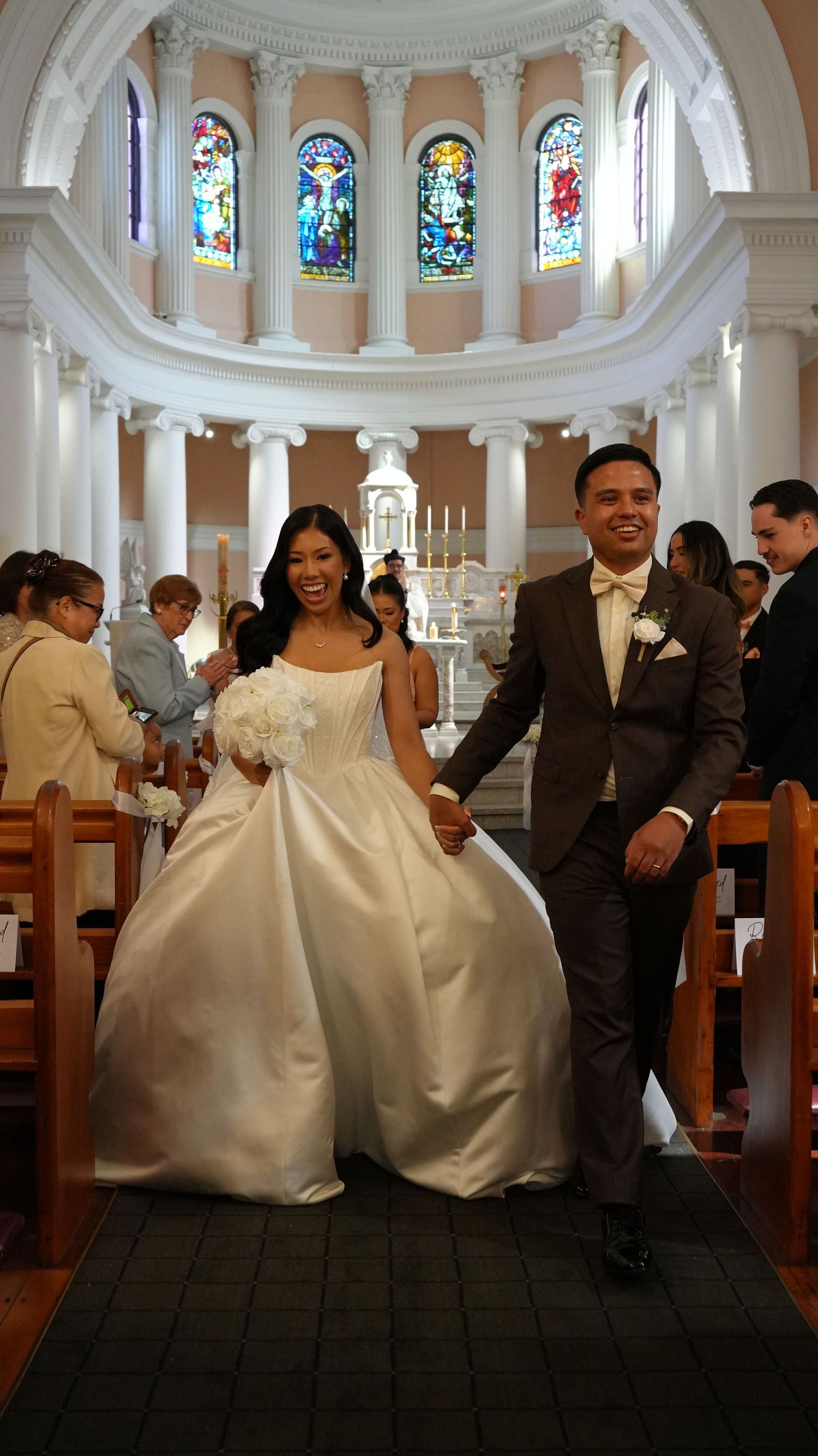 Smiling newlyweds walk down church aisle, bride in white ball gown, groom in brown suit, stained glass windows behind.