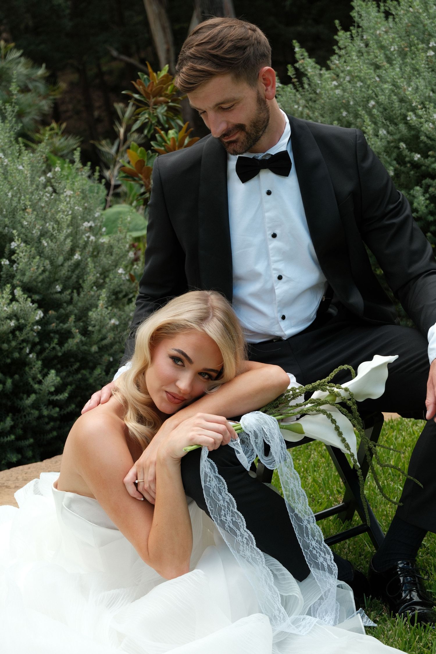 A romantic wedding photo of a couple resting on a garden bench surrounded by greenery.