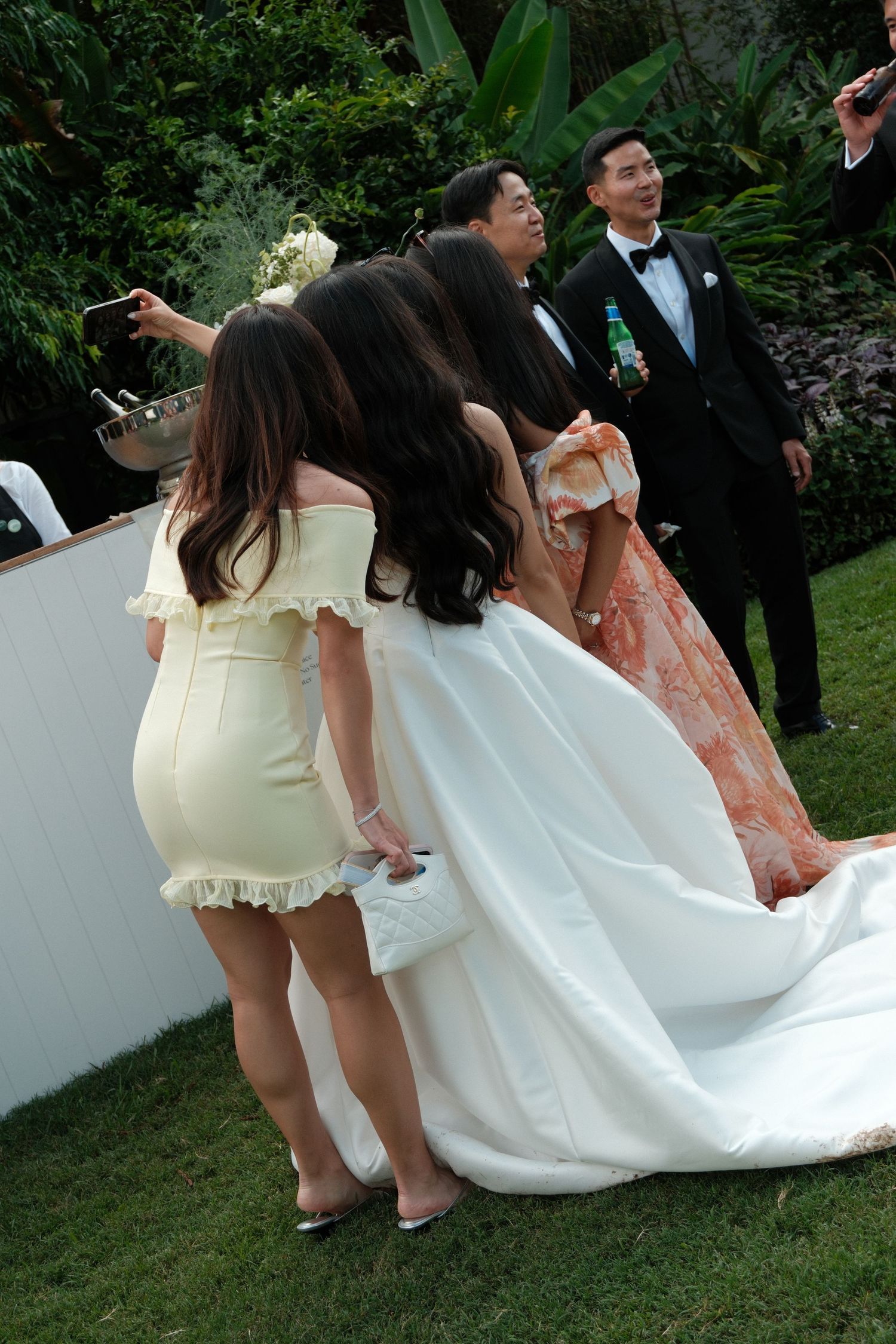 Young children in formal attire lift and play with a white wedding dress train on a grassy lawn during an outdoor ceremony.