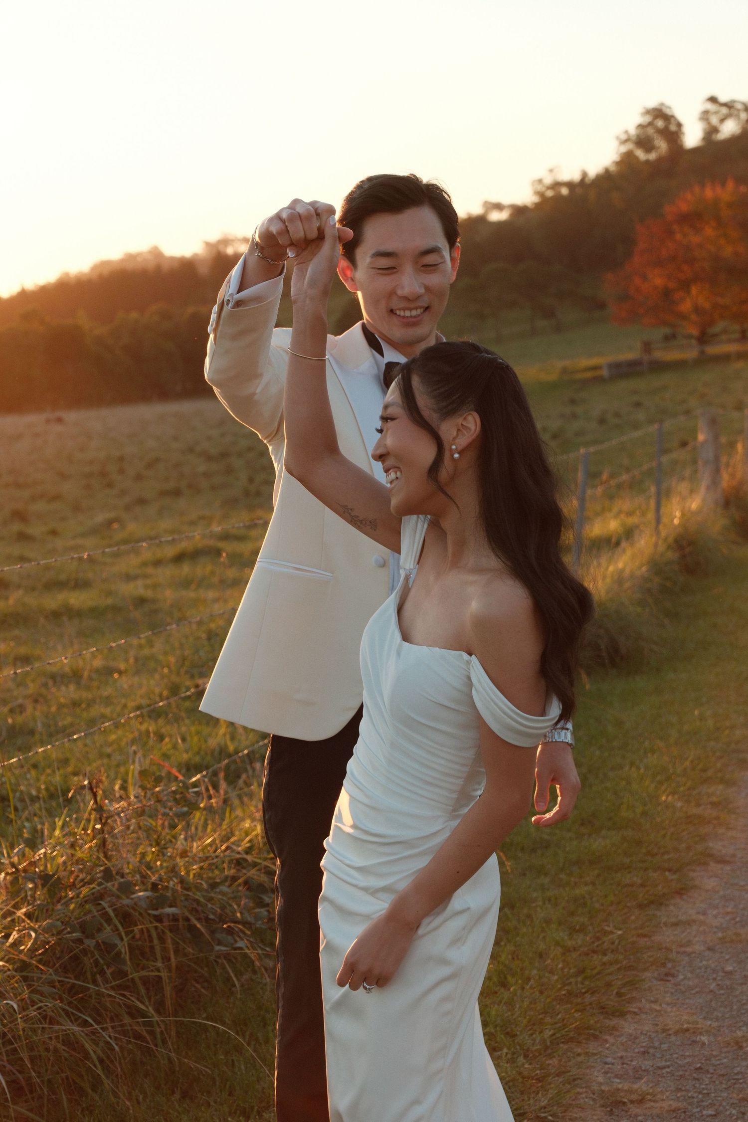 Couple in wedding attire dancing at golden hour sunset on a scenic countryside path, bride in white gown, groom in white tuxedo.