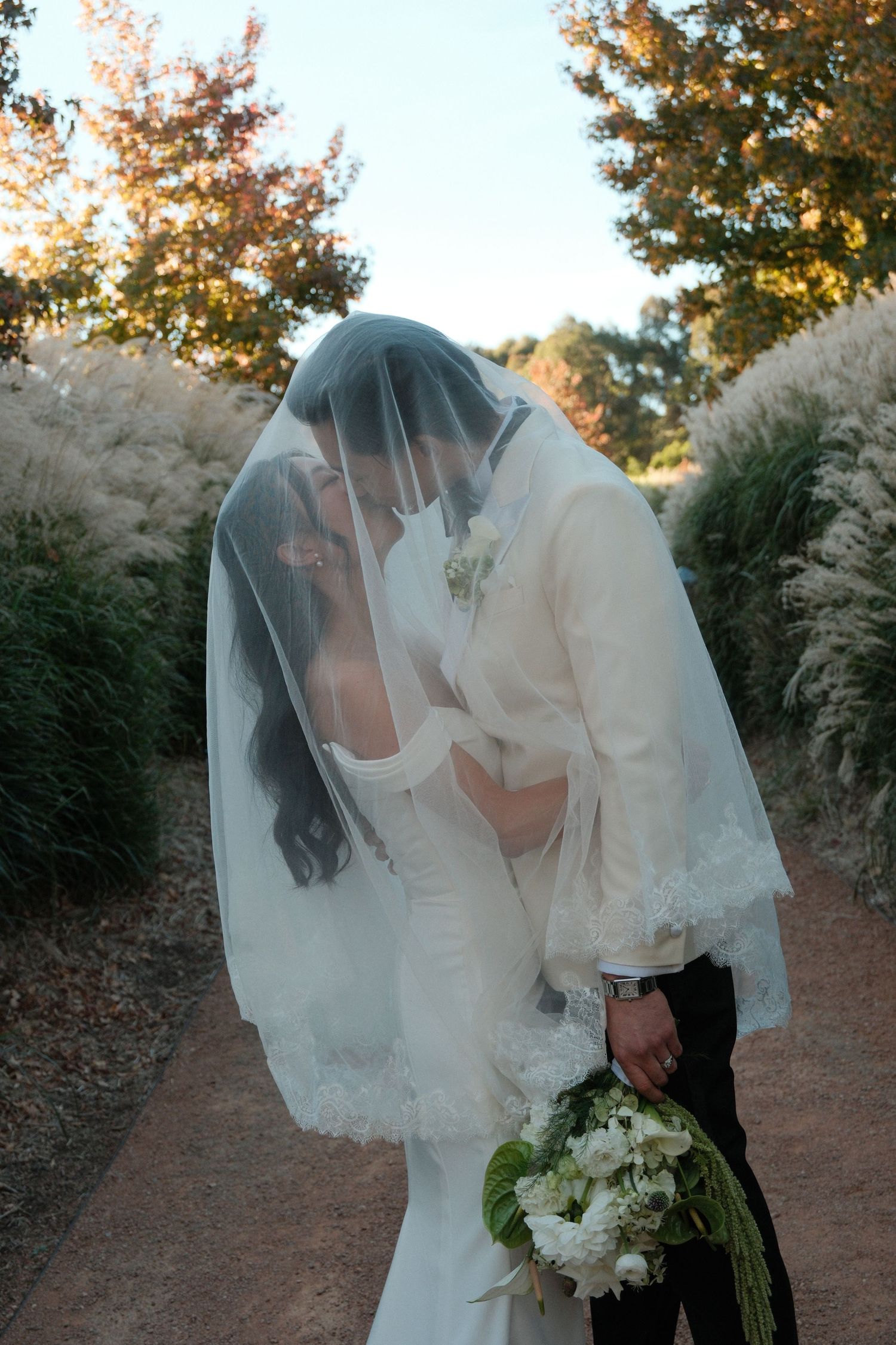 Bride and groom share a romantic kiss under a lace-trimmed wedding veil outdoors surrounded by autumn foliage.