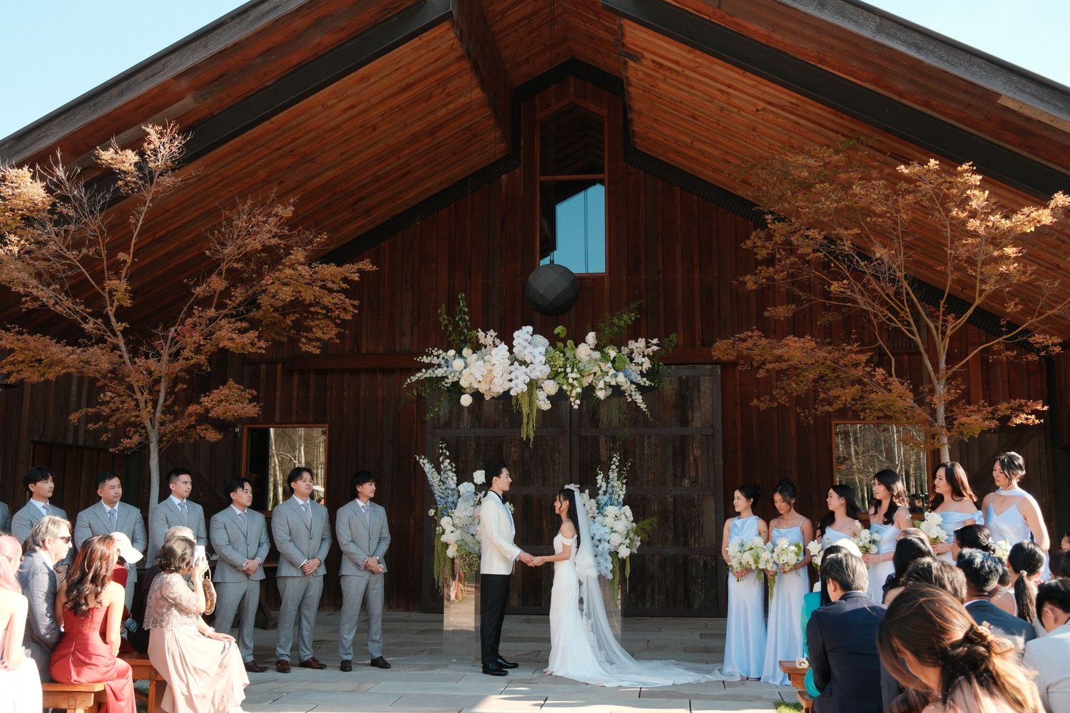Outdoor barn wedding ceremony with bride and groom at floral arch, wedding party in gray suits and blue dresses.