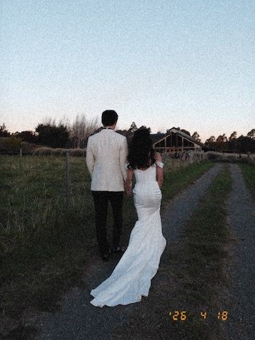 A bride in a white gown and groom in a tan suit walk hand in hand down a rural path at dusk.