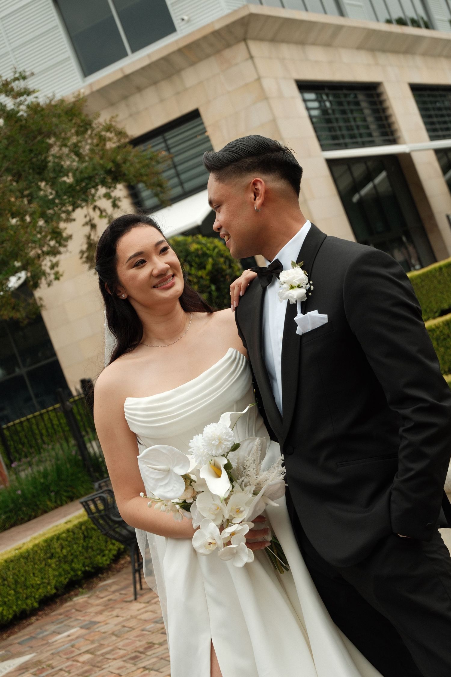 A couple shares a tender moment in formal wedding attire on a stone paved walkway outside an elegant building.