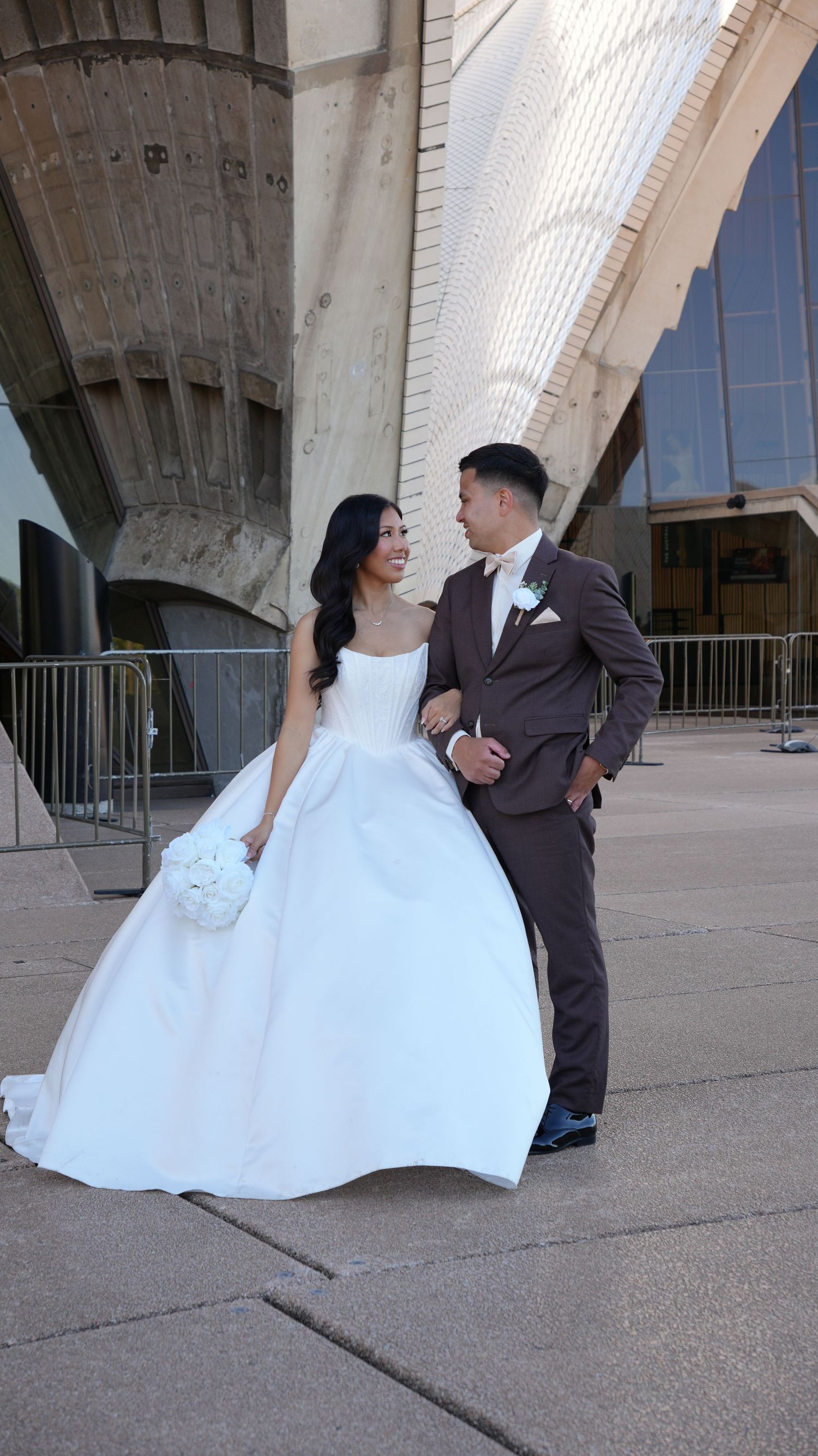 Bride in white ball gown holding bouquet stands with groom in brown suit outside modern architectural venue.