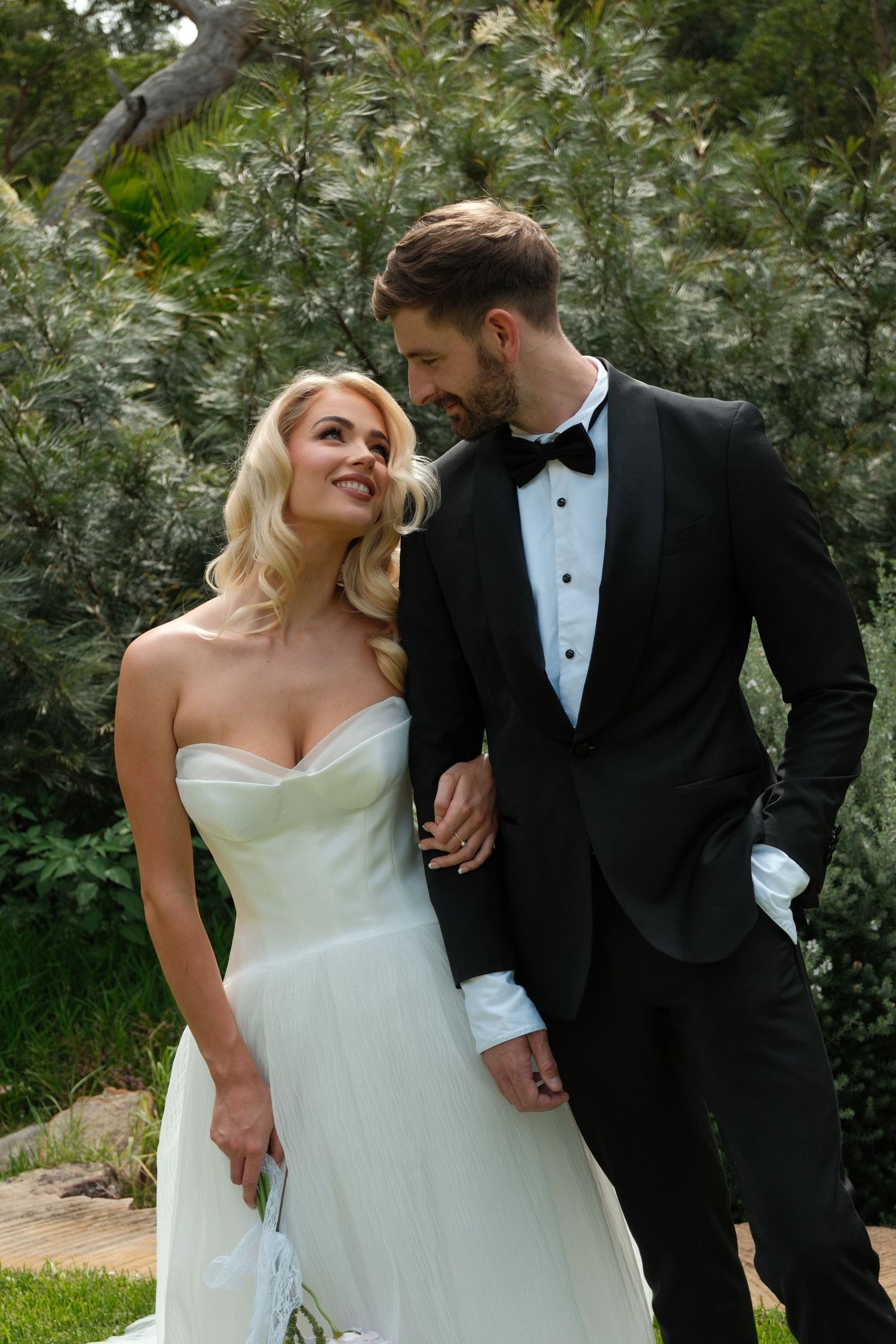 A bride in a white strapless gown and groom in black tuxedo share a romantic moment outdoors among lush greenery.