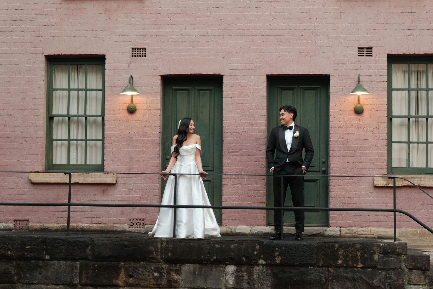 Bride and groom stand apart on a stone ledge before a pink brick building with green doors.